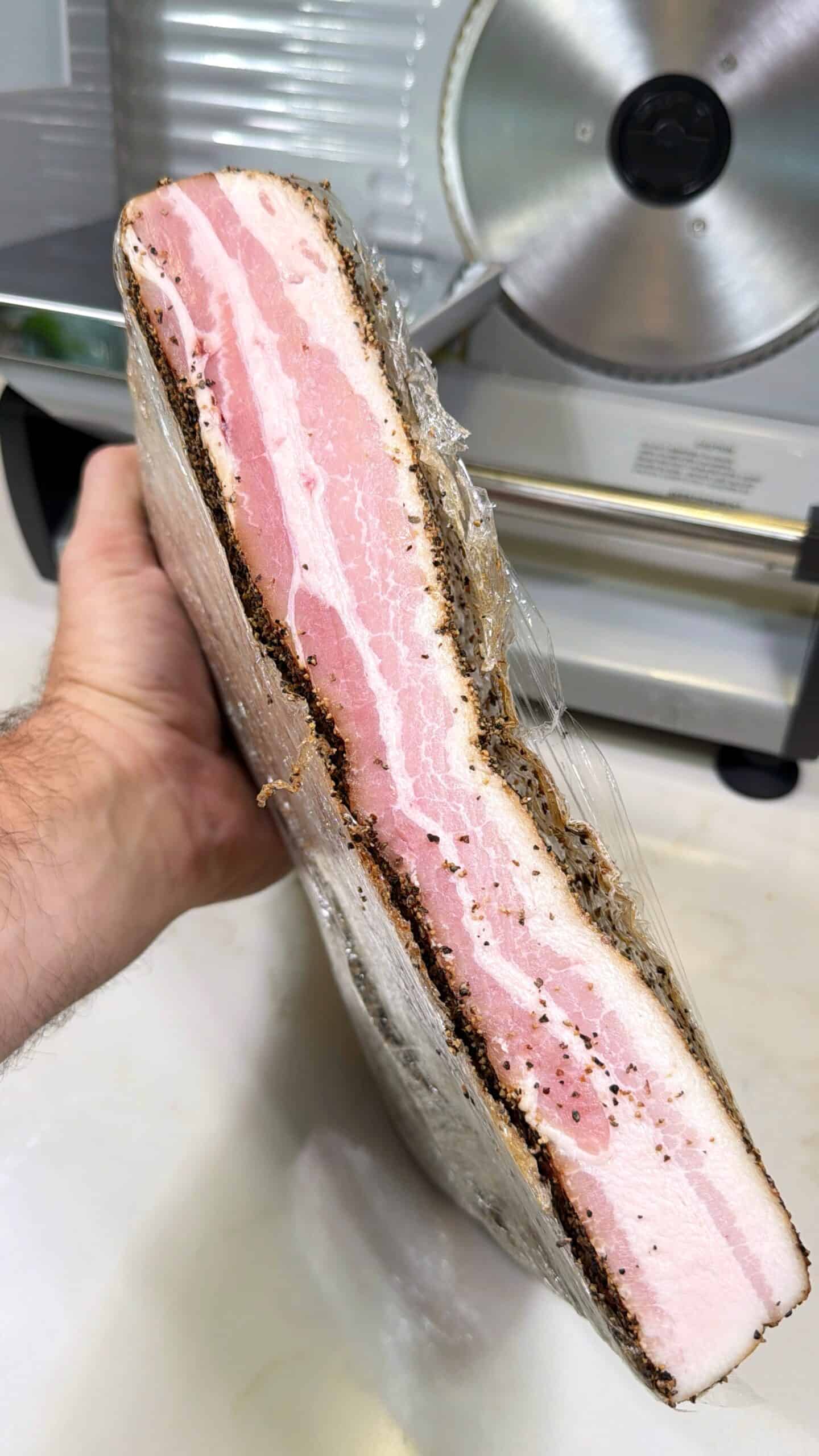 A hand holds a thick slab of uncooked pastrami bacon with a peppered edge, wrapped in plastic, in front of a meat slicer on a kitchen counter.