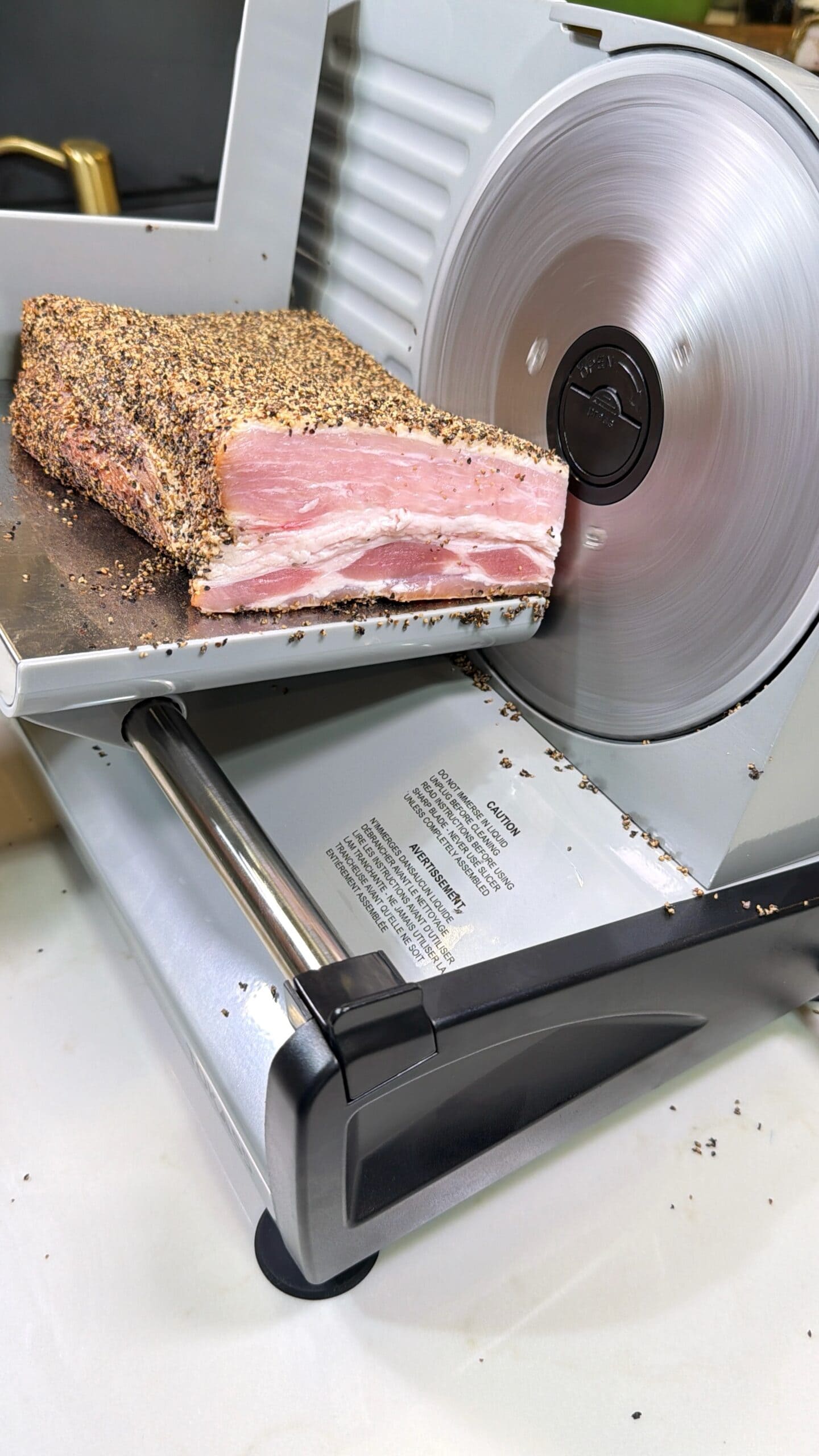 A close-up of a meat slicer cutting through a slab of pepper-crusted bacon, with seasoning scattered on the slicer and countertop.