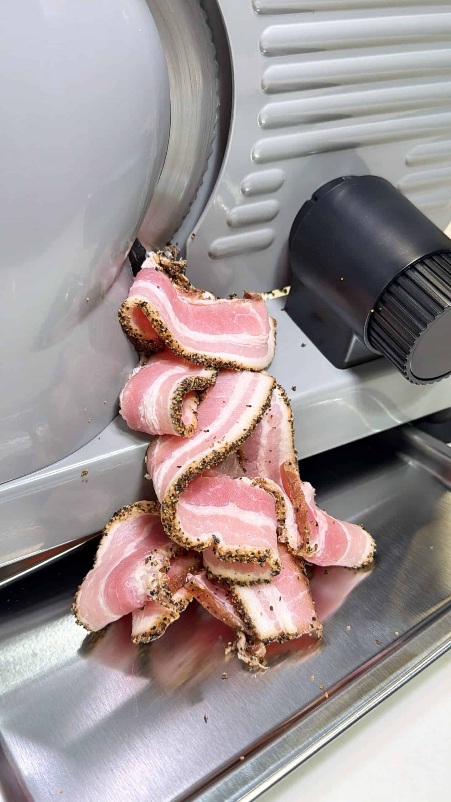 Several slices of pepper-crusted bacon are being cut by an electric meat slicer, with the freshly sliced pieces piling up on the metal tray below.