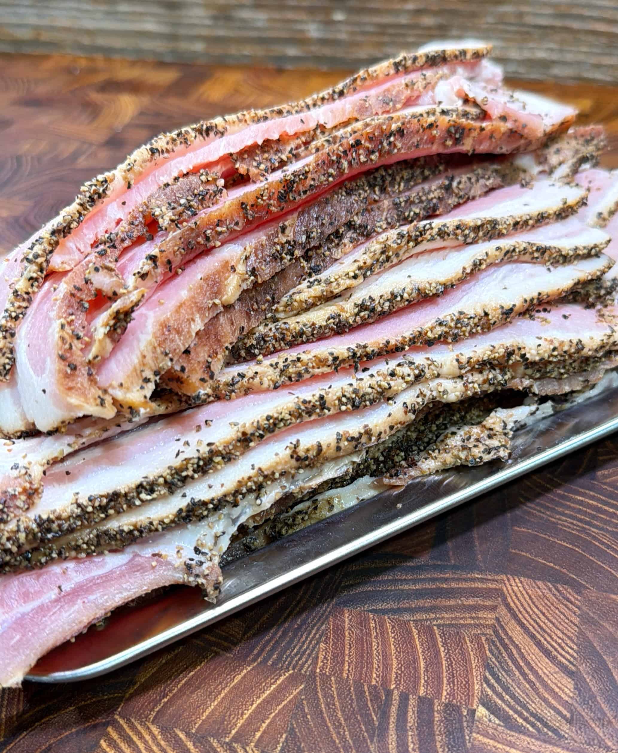 Several uncooked, thick-cut strips of pepper-crusted bacon are stacked on a metal tray, placed on a wooden surface with a geometric pattern.