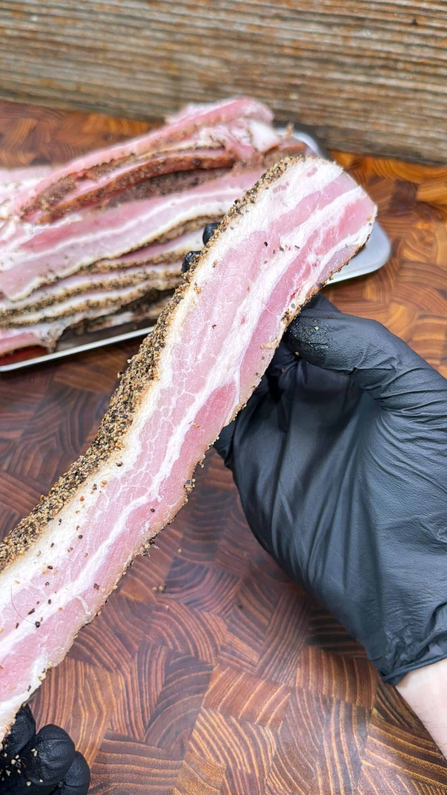 A hand in a black glove holds a thick, uncooked slice of peppered bacon over a wooden surface, with more slices stacked on a plate in the background.
