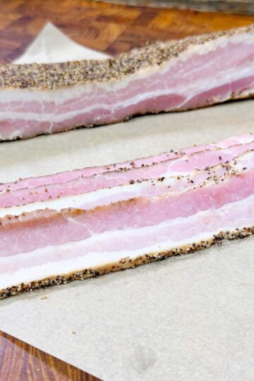 Two thick slices of uncooked, pepper-crusted bacon with visible layers of pink meat and white fat are placed on a piece of brown parchment paper on a wooden surface.