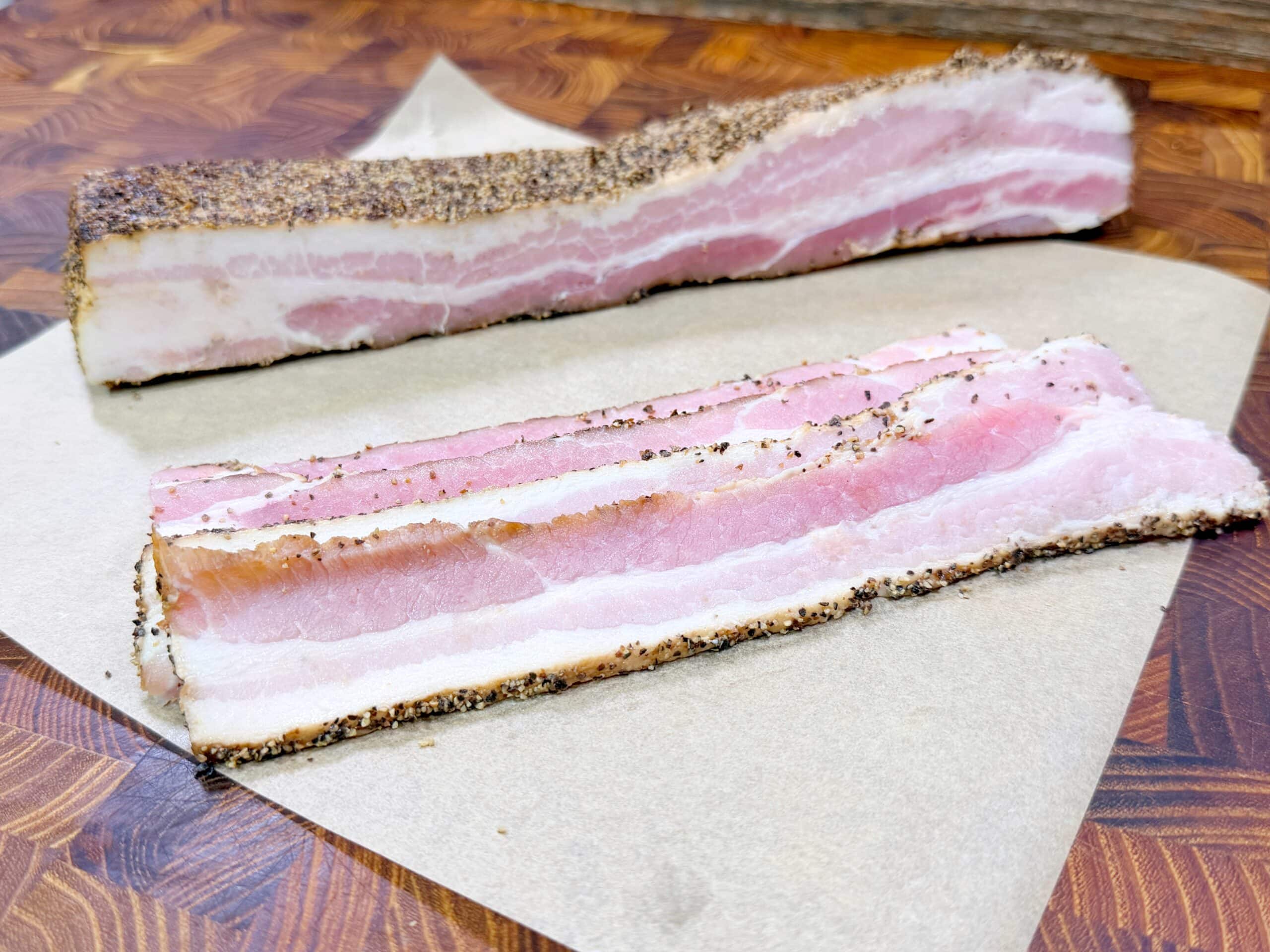 Two thick slices of uncooked, pepper-crusted bacon with visible layers of pink meat and white fat are placed on a piece of brown parchment paper on a wooden surface.