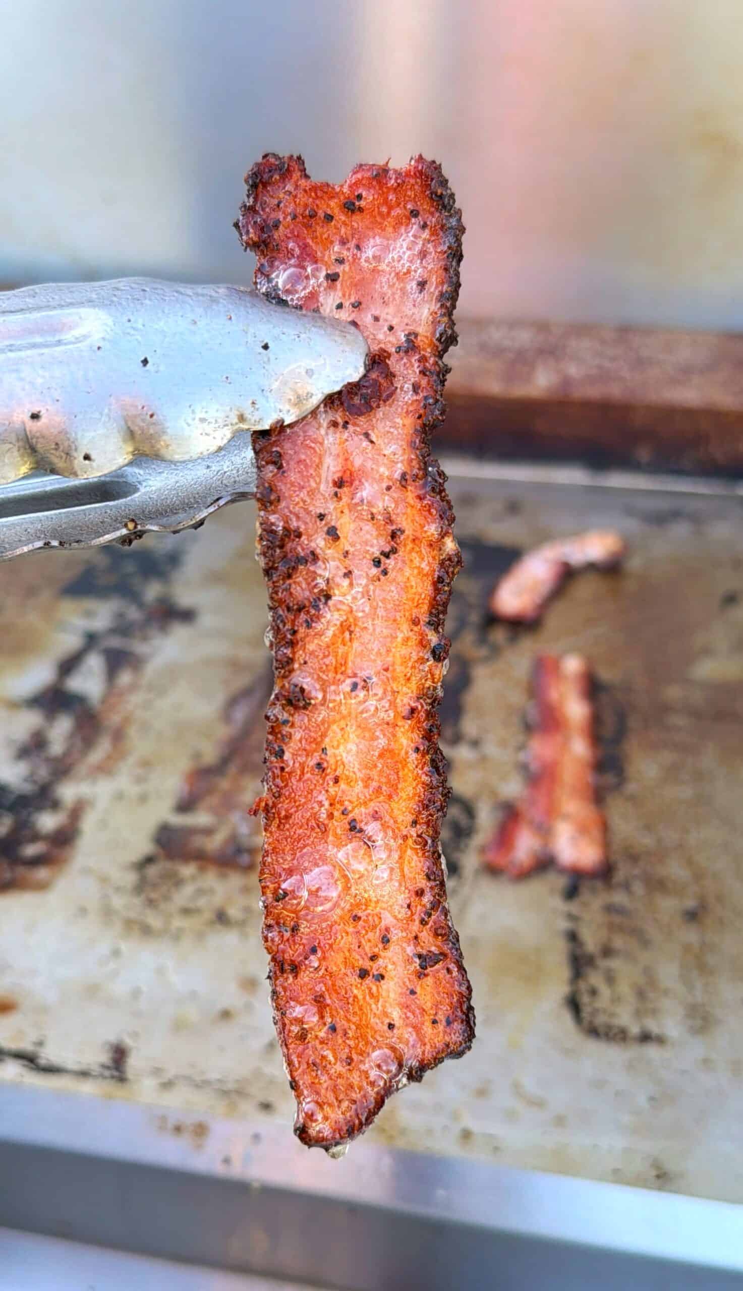 A close-up of metal tongs holding a crispy, peppered strip of bacon, with a griddle and more bacon strips blurred in the background.