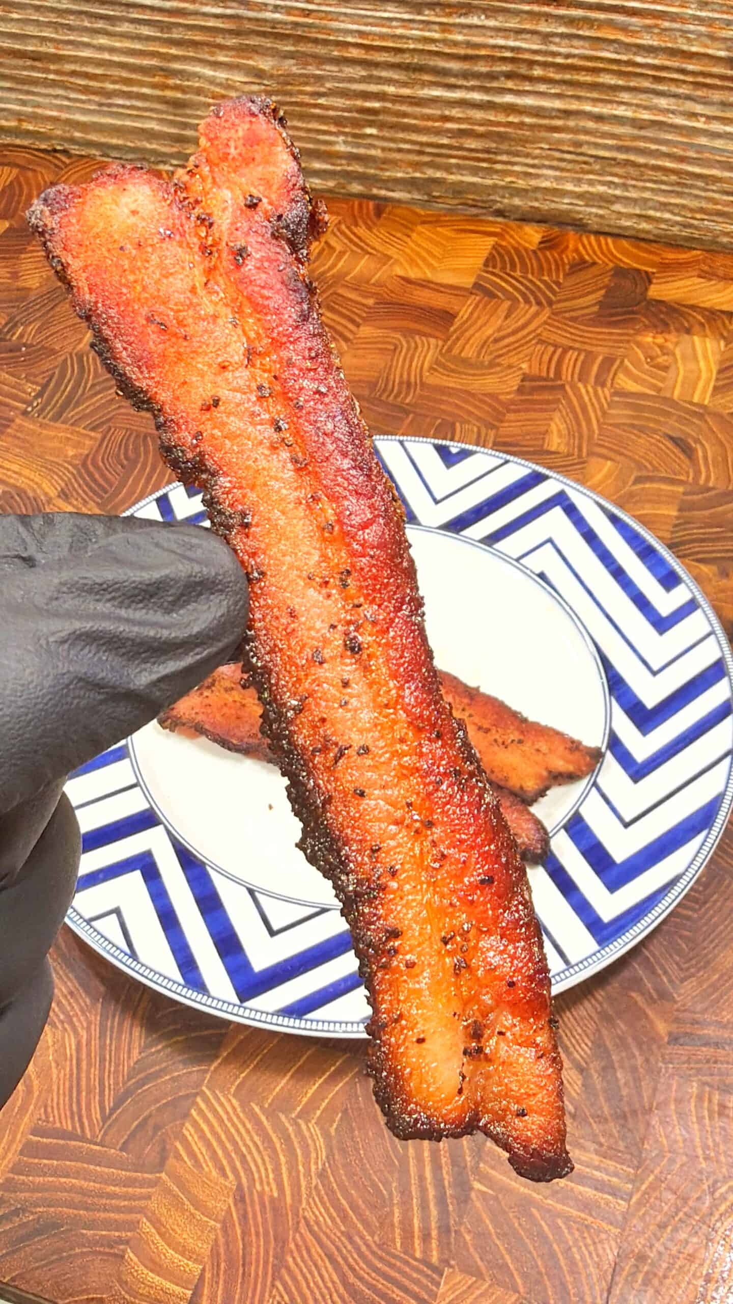A gloved hand holds a crispy, seasoned strip of bacon over a plate with more bacon strips, on a wooden surface with a zigzag-patterned plate.