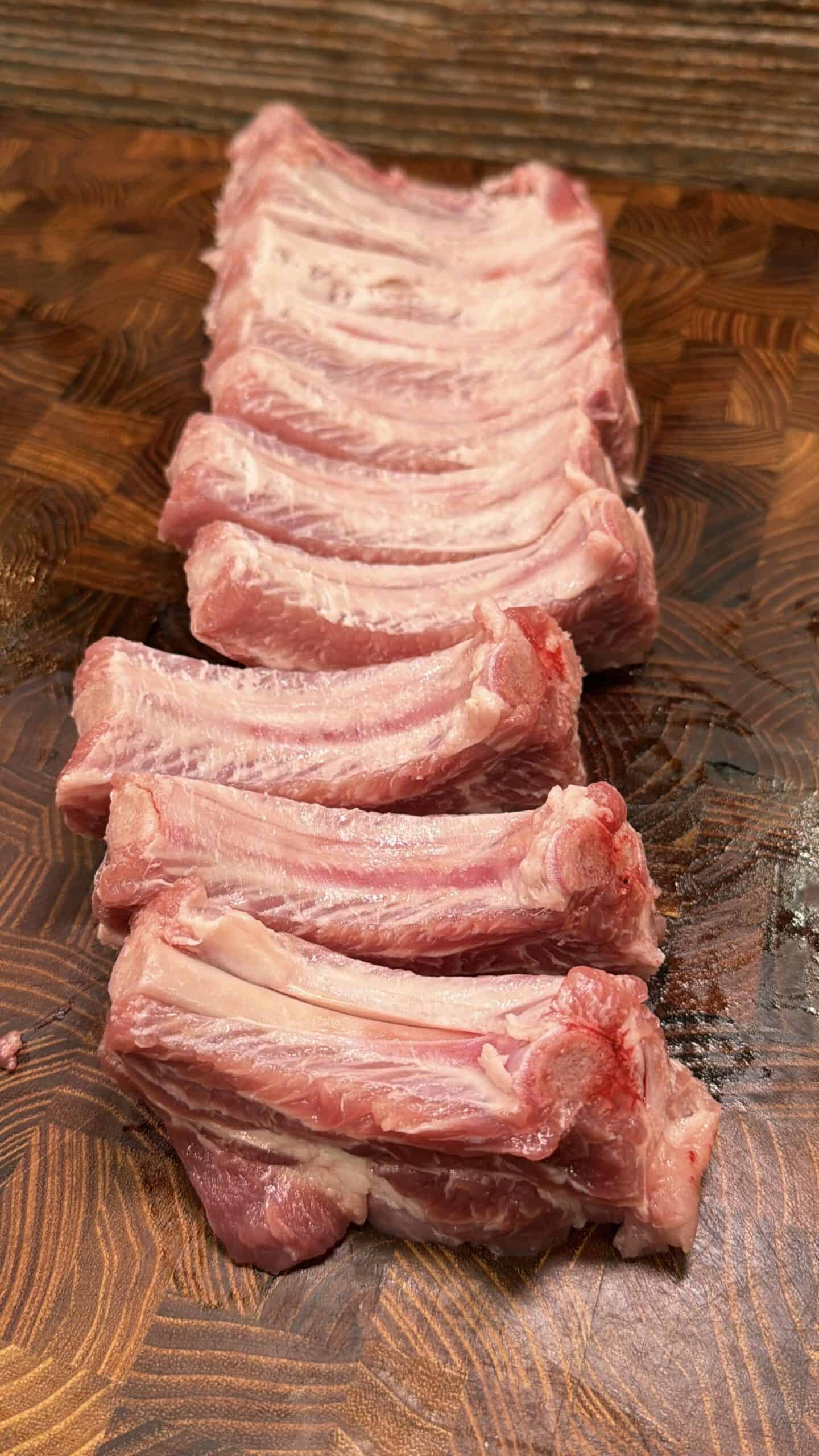 Raw pork ribs arranged in a neat row on a wooden cutting board with a patterned surface. The ribs are uncooked and show visible marbling and a bit of red near the edges.