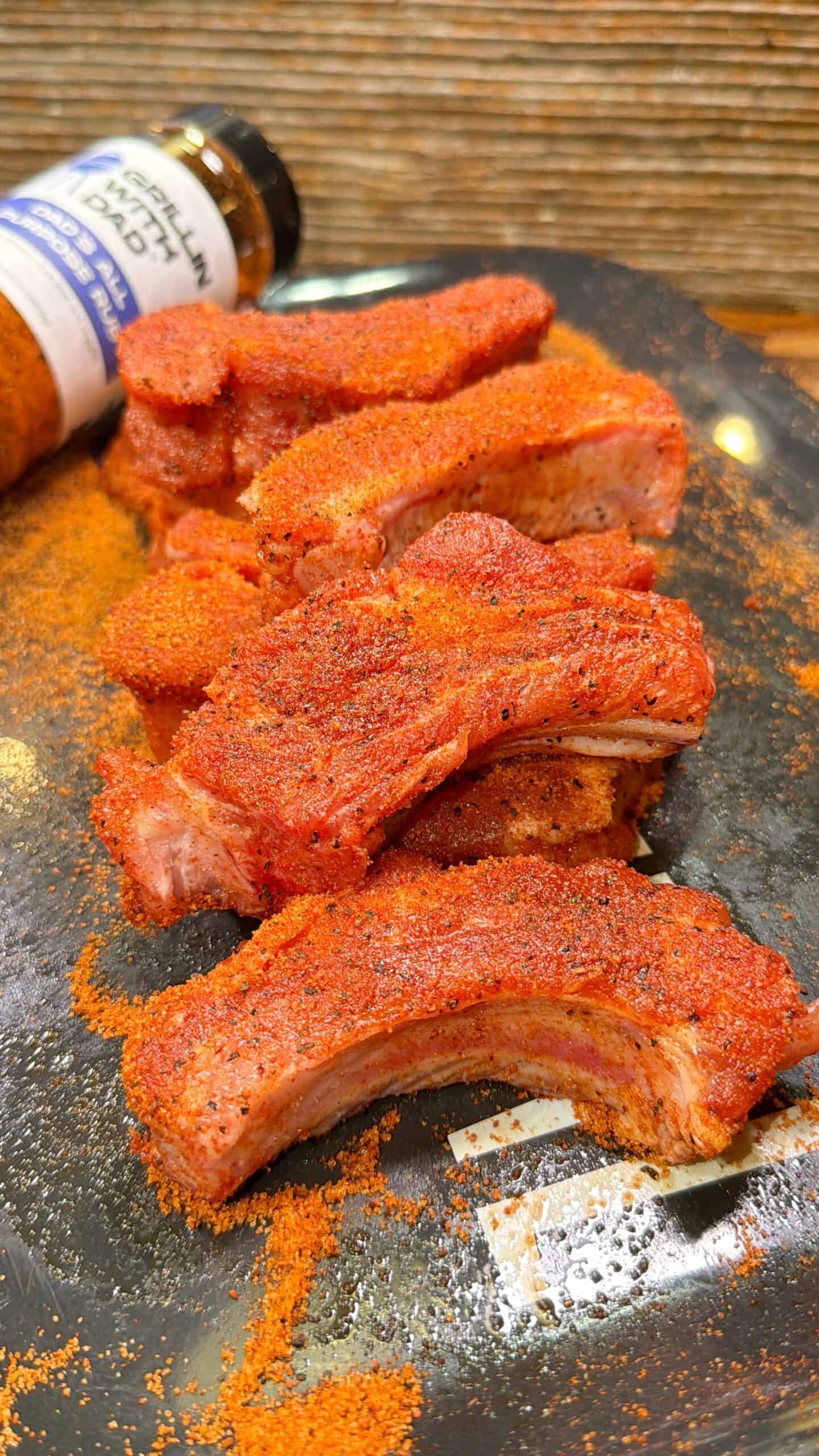Five seasoned pork ribs are arranged on a dark plate, generously coated with a red, spicy dry rub. In the background, there is a bottle of seasoning, partially visible and tipped over.
