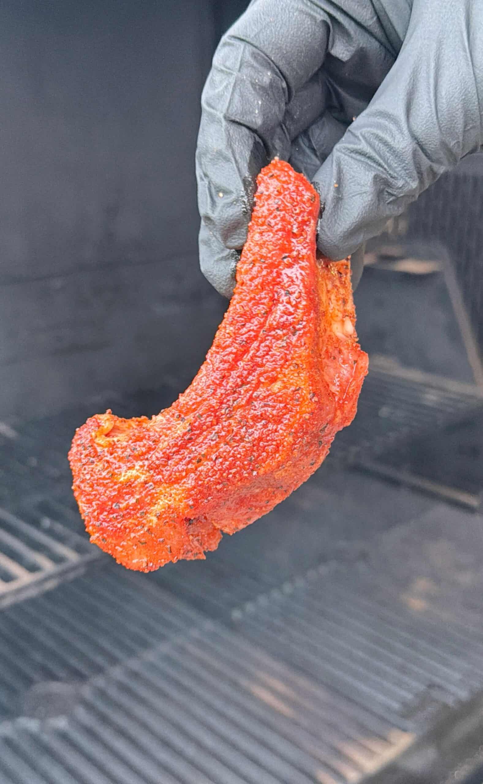 A gloved hand holds a seasoned, uncooked pork rib in front of a barbecue grill, ready for smoking or grilling.