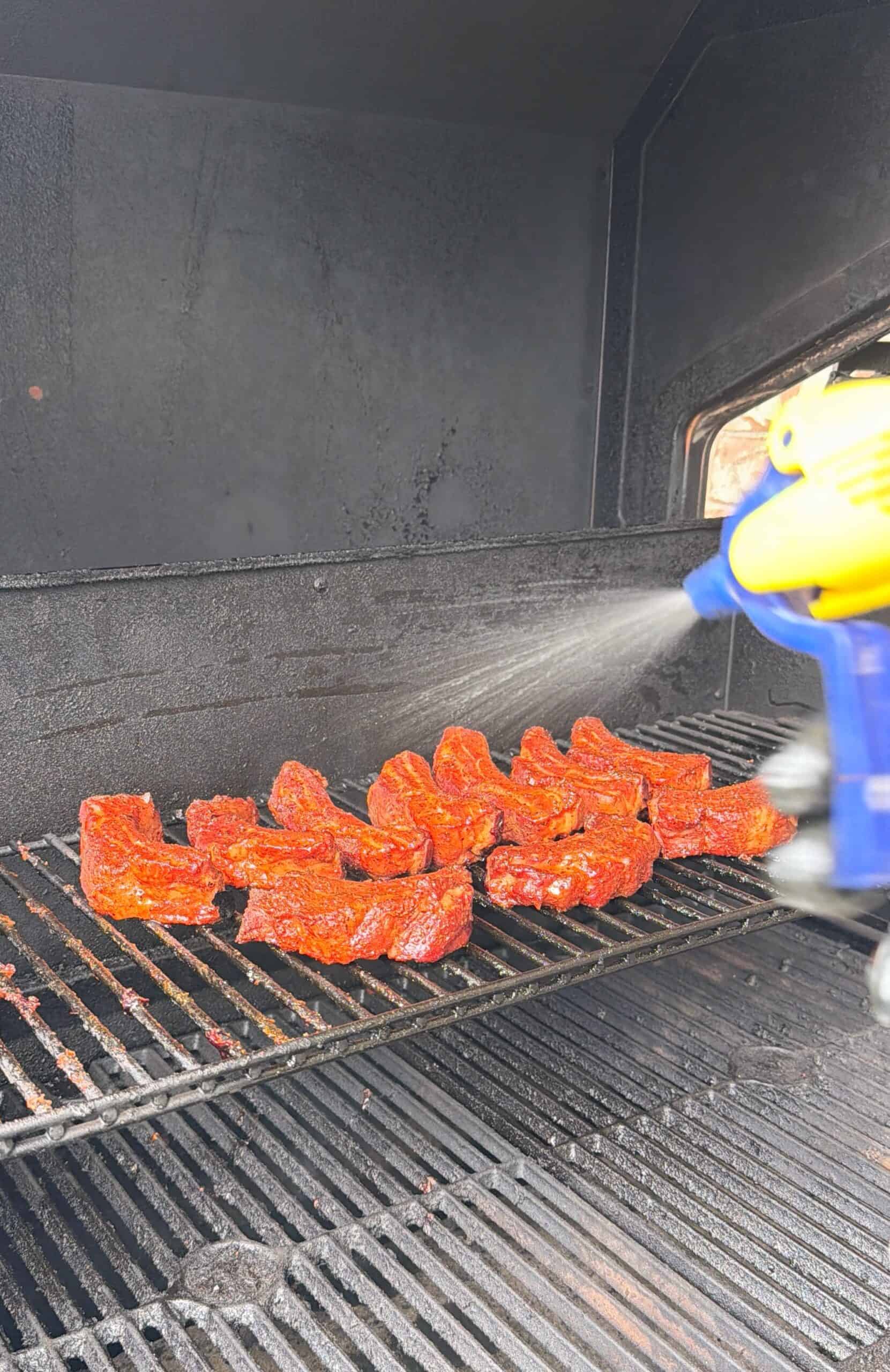 Strips of marinated meat are arranged on a grill inside a smoker while a blue and yellow spray bottle mists them, keeping the meat moist during cooking.