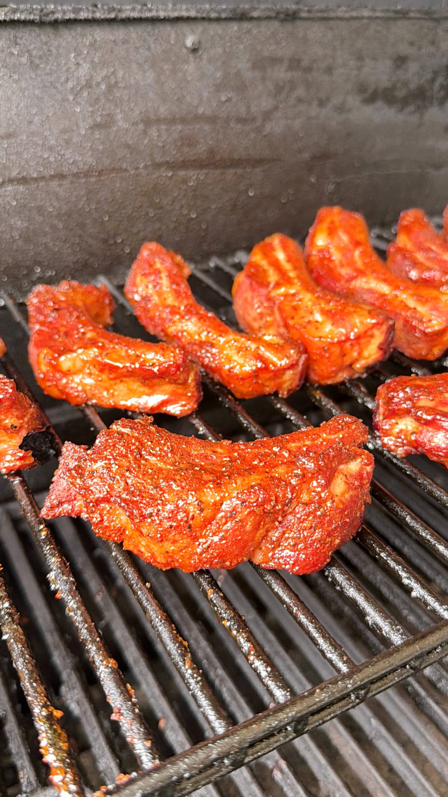 Several pieces of marinated meat with a red seasoning are cooking on a metal grill inside a smoker or barbecue. The meat is sizzling and appears to be in the process of being smoked or grilled.