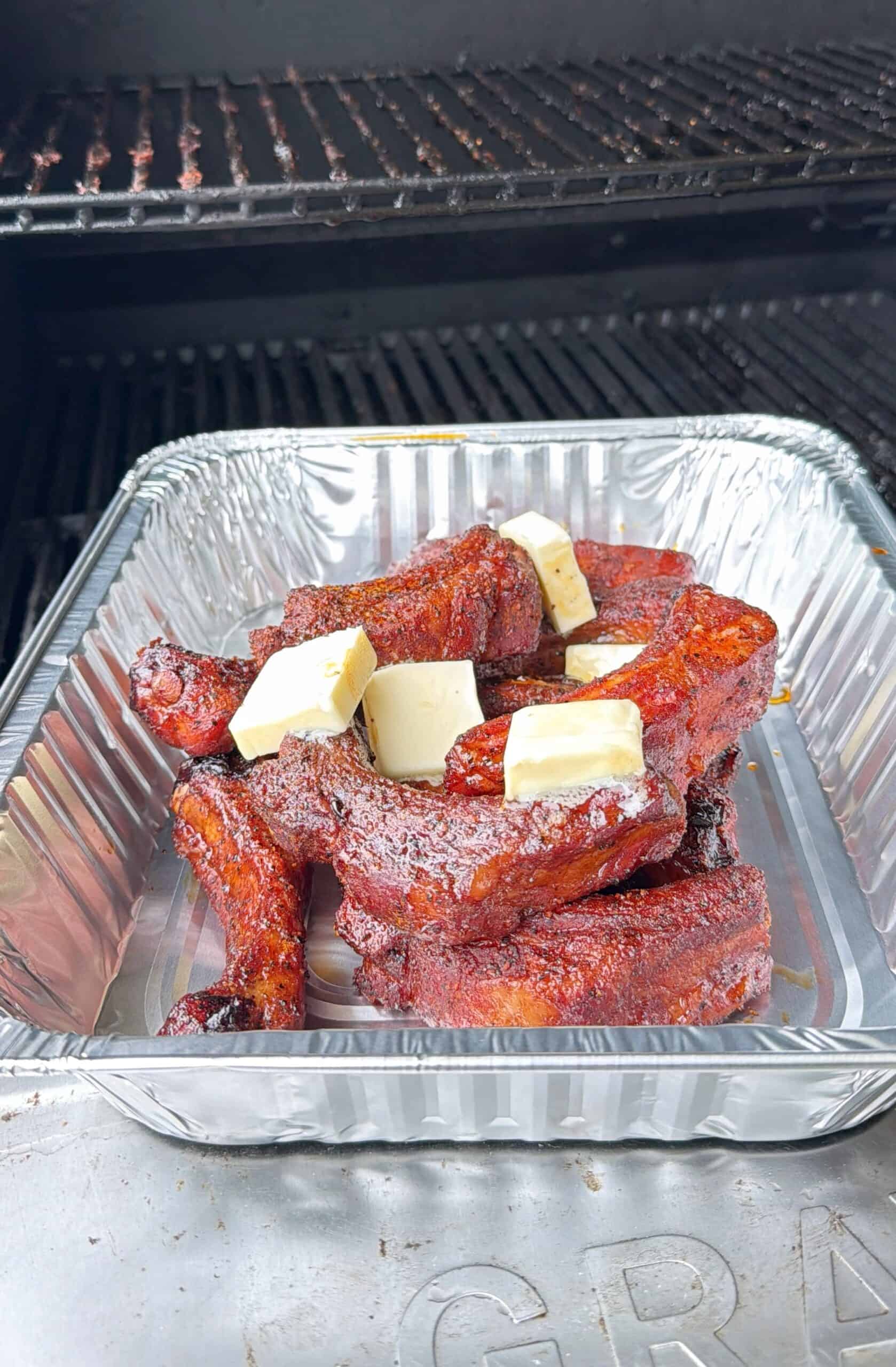 A foil tray filled with smoked pork ribs topped with several cubes of butter sits on an outdoor grill. The ribs are coated in seasoning and have a reddish-brown, caramelized appearance.