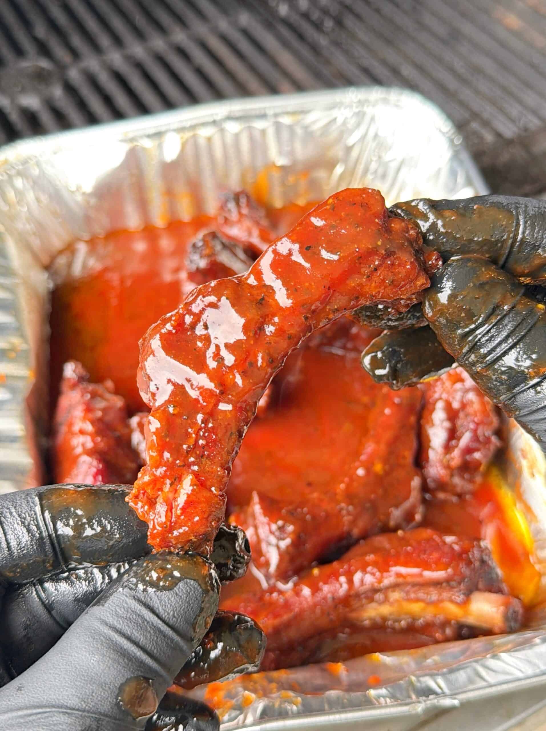 Gloved hands hold a saucy, barbecued rib above a tray filled with more ribs and red barbecue sauce, near a grill.  Recipe is for Sweet Tea Party Ribs