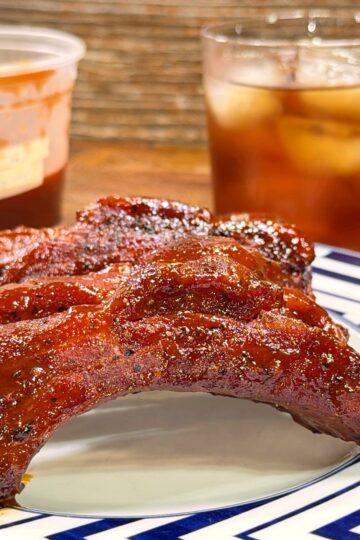 A plate with barbecue ribs sits on a geometric-patterned plate, with a glass of iced drink and a plastic container of barbecue sauce in the background on a wooden surface.