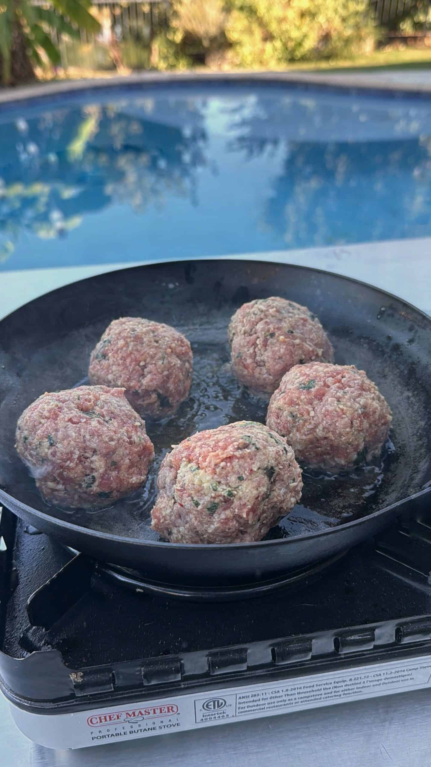 Five raw meatballs are cooking in a black skillet on an outdoor stove, with a swimming pool and greenery visible in the background. Steam rises from the hot pan.