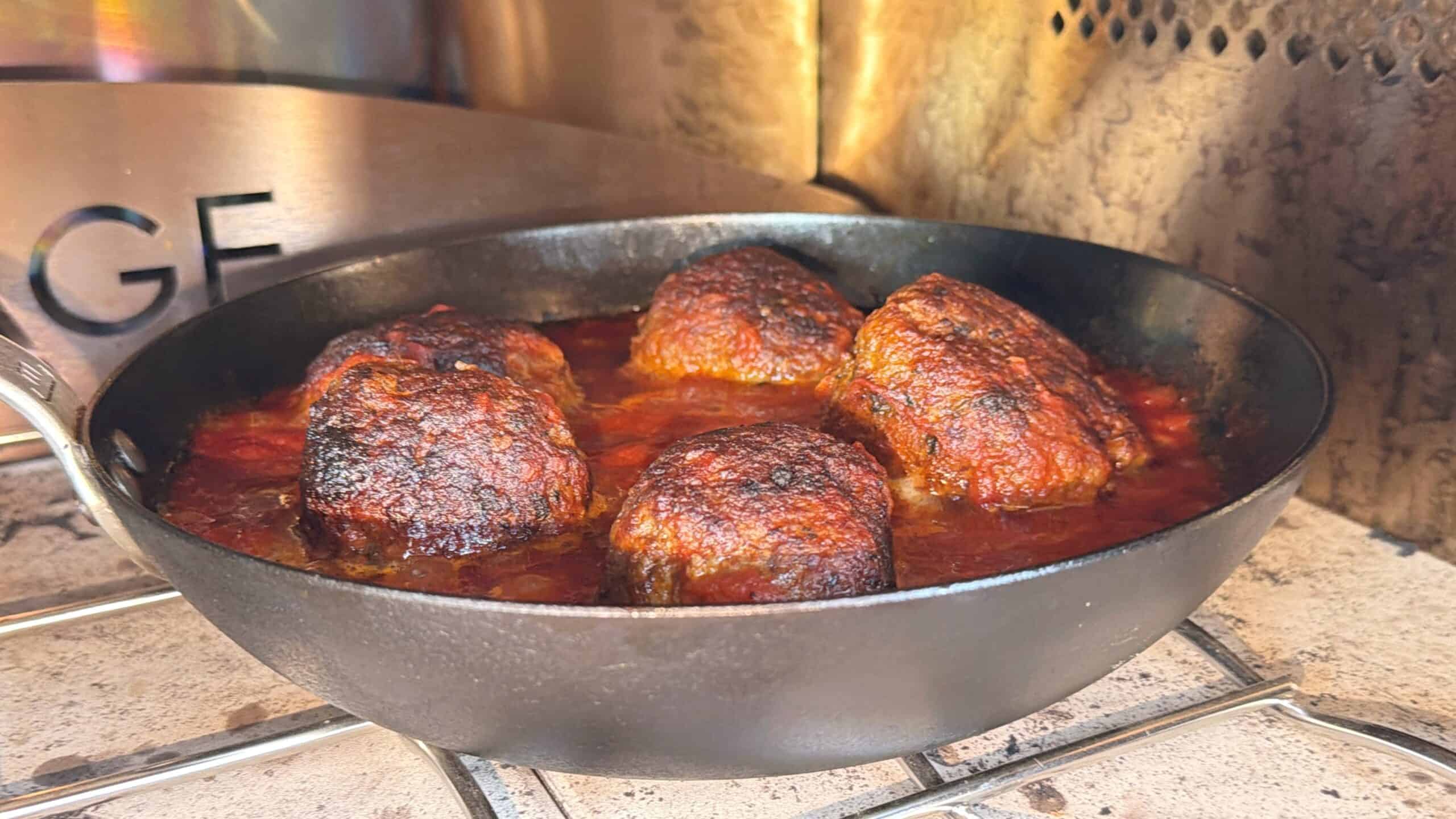 Five large meatballs simmer in a cast iron skillet filled with red tomato sauce. The meatballs have a crispy, browned exterior, and the pan sits inside an oven with metal walls and a rack visible.