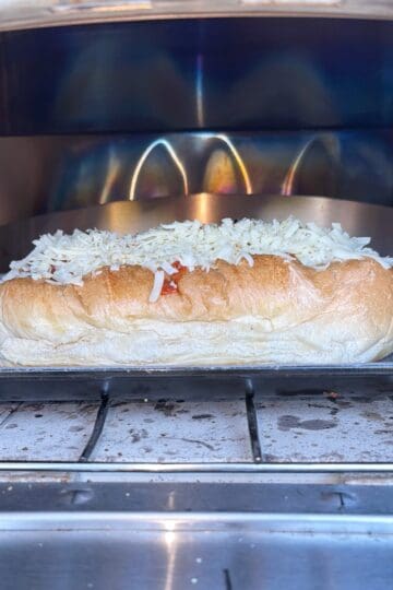 A sub sandwich topped with shredded cheese is on a wire rack inside an oven, being toasted.