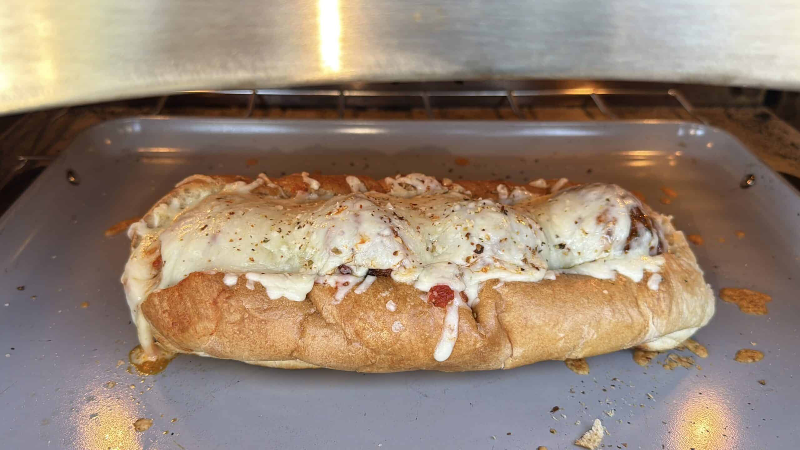 A toasted sub sandwich on a baking tray in the oven, topped with melted mozzarella cheese, tomato sauce, and sprinkled herbs.