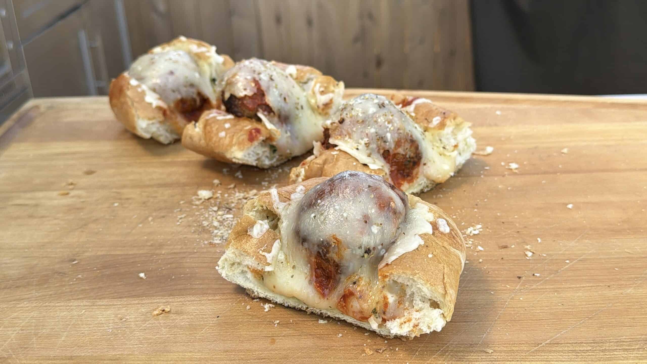 A wooden cutting board with a sliced meatball party sandwich topped with melted cheese and tomato sauce, with crumbs scattered around.