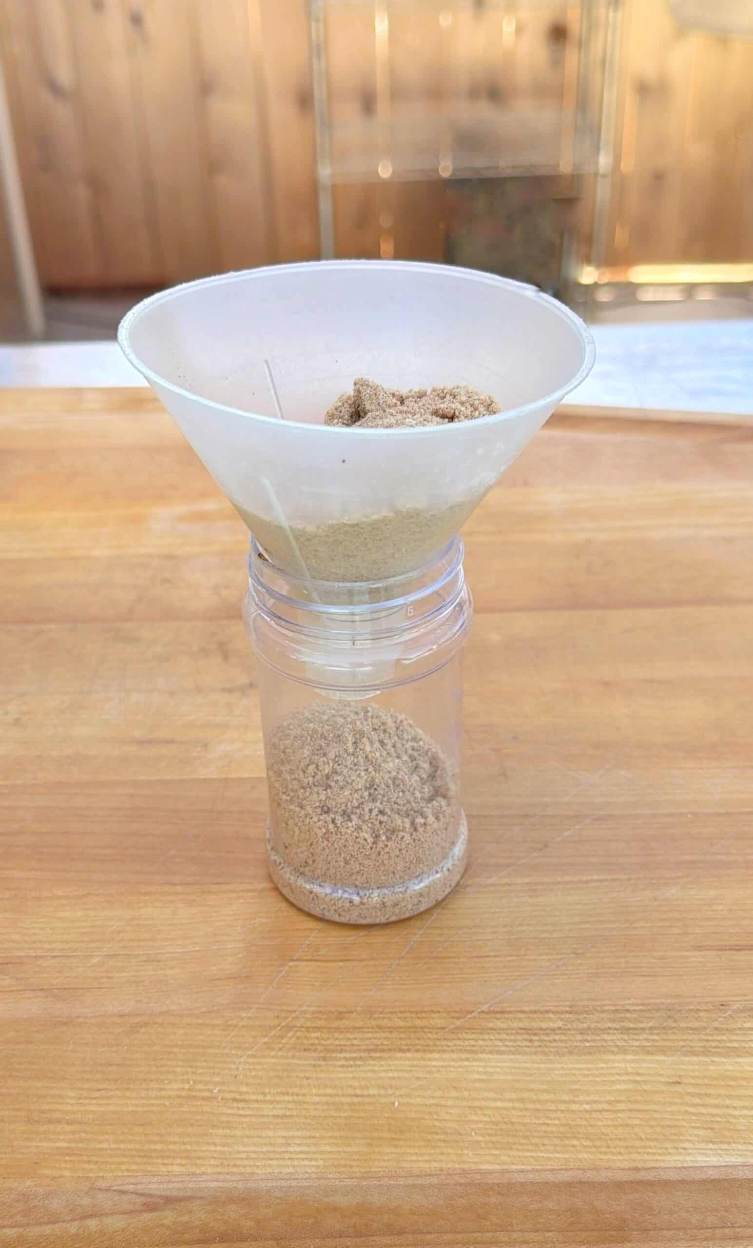 A white funnel filled with brown sugar sits atop a clear plastic container, with some brown sugar collected inside, all placed on a wooden surface.