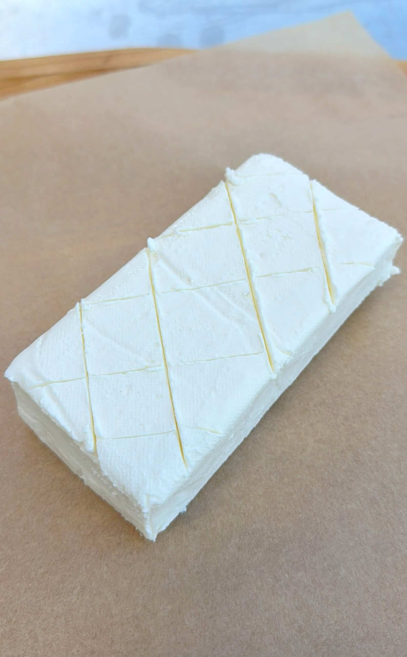 A rectangular block of cream cheese with a smooth, creamy surface and faint diagonal lines etched on top, placed on a sheet of brown parchment paper.