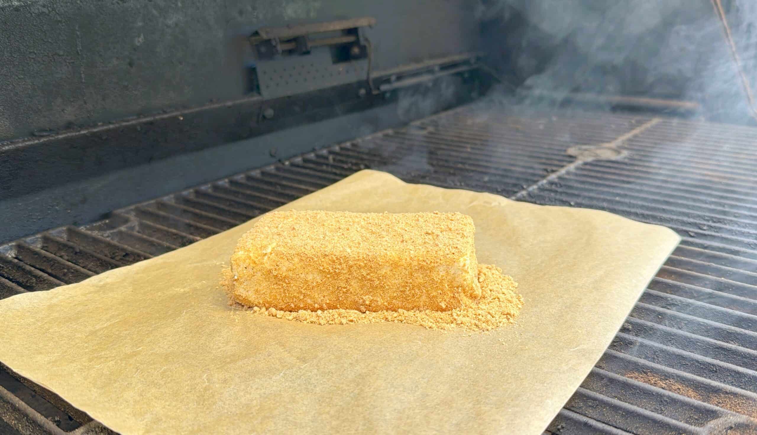 A rectangular, crumb-coated block sits on parchment paper on a grill with smoke rising, suggesting it is being smoked or cooked. The grill grates and part of the grill’s interior are visible.