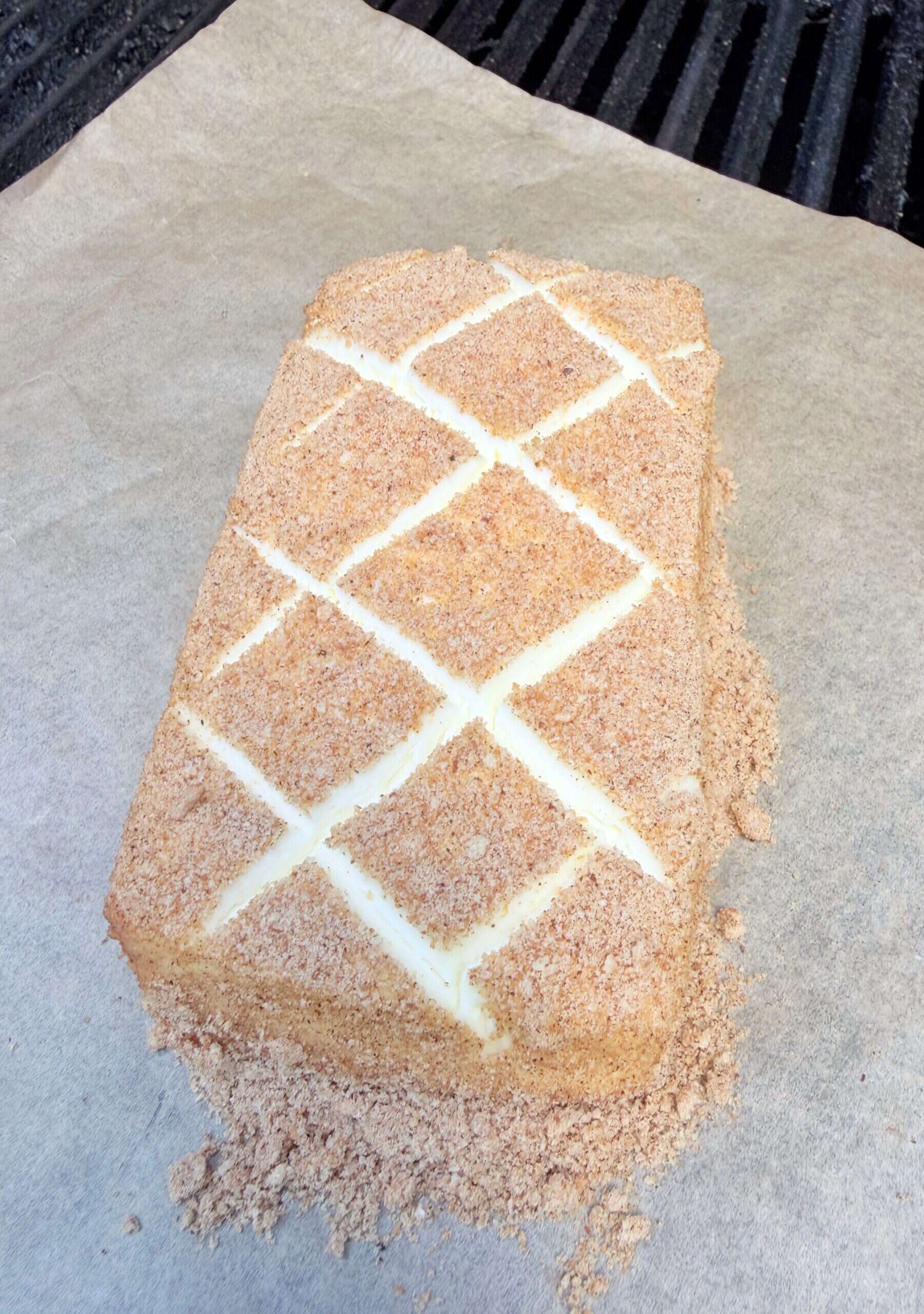 A rectangular block of cinnamon toast crunch smoked cream cheese coated with breadcrumbs sits on parchment paper, featuring a diamond pattern scored on top.