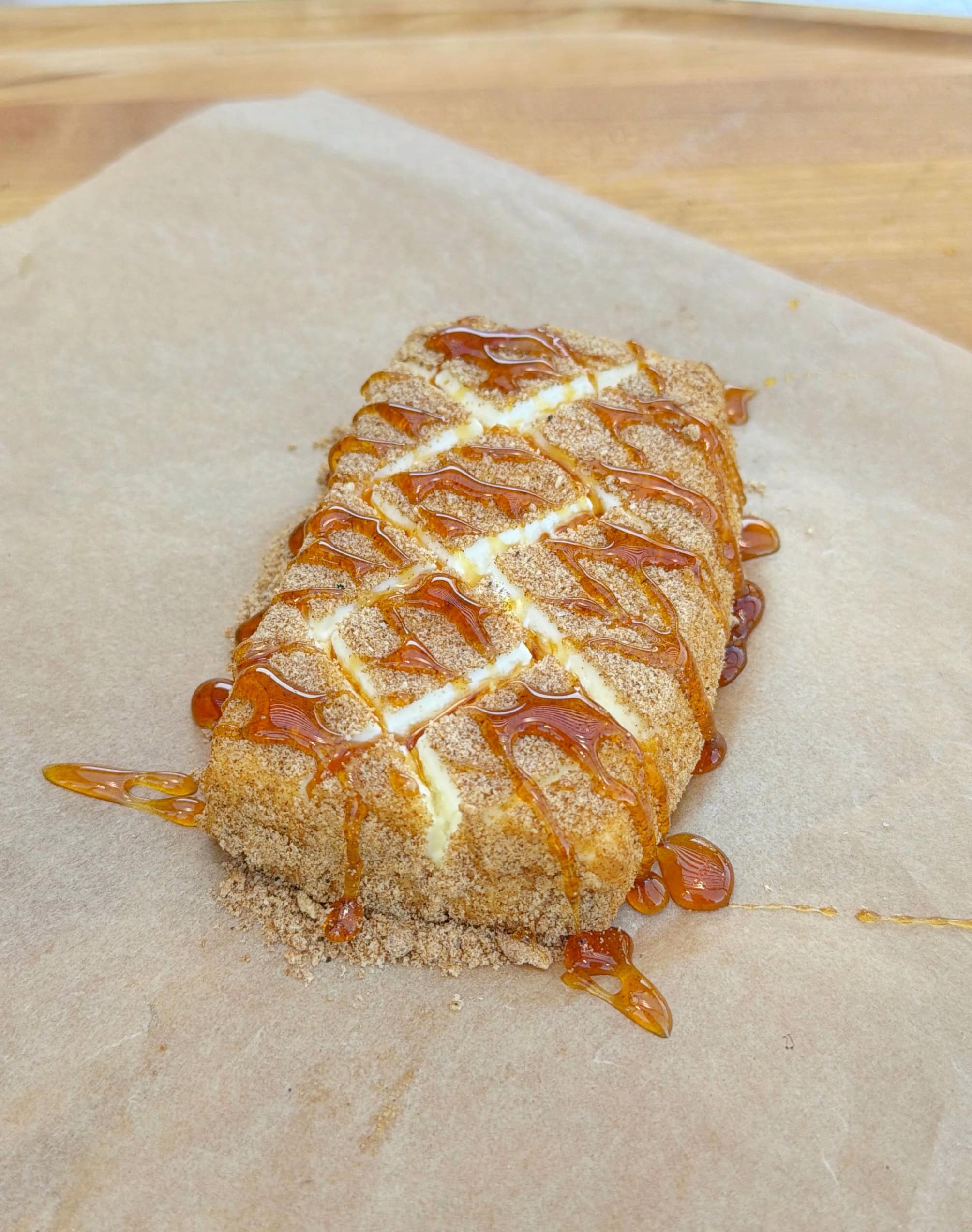 A rectangular pastry dusted with brown sugar, drizzled with caramel sauce, and scored with diagonal lines, sits on brown parchment paper on a wooden surface.