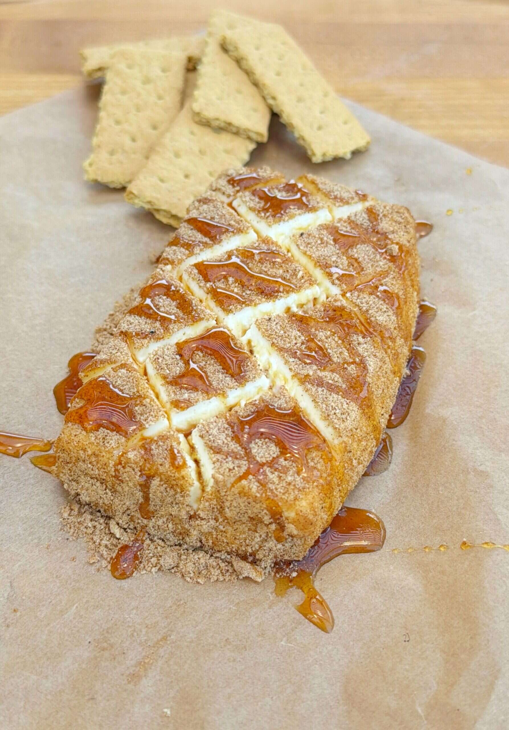 A rectangular cheesecake covered in brown sugar and caramel sauce, sliced into pieces, sits on parchment paper with several broken graham crackers behind it on a wooden surface.