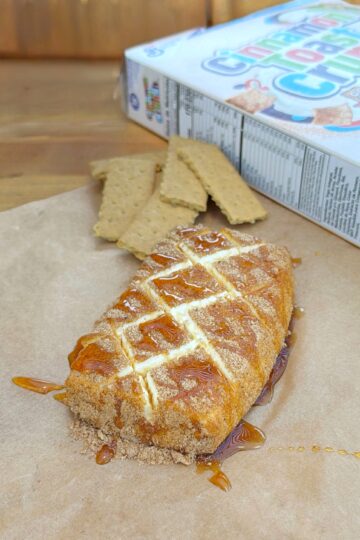 A golden-brown pastry topped with white icing and caramel sauce sits on parchment paper, with graham cracker pieces and a partially visible cereal box in the background.