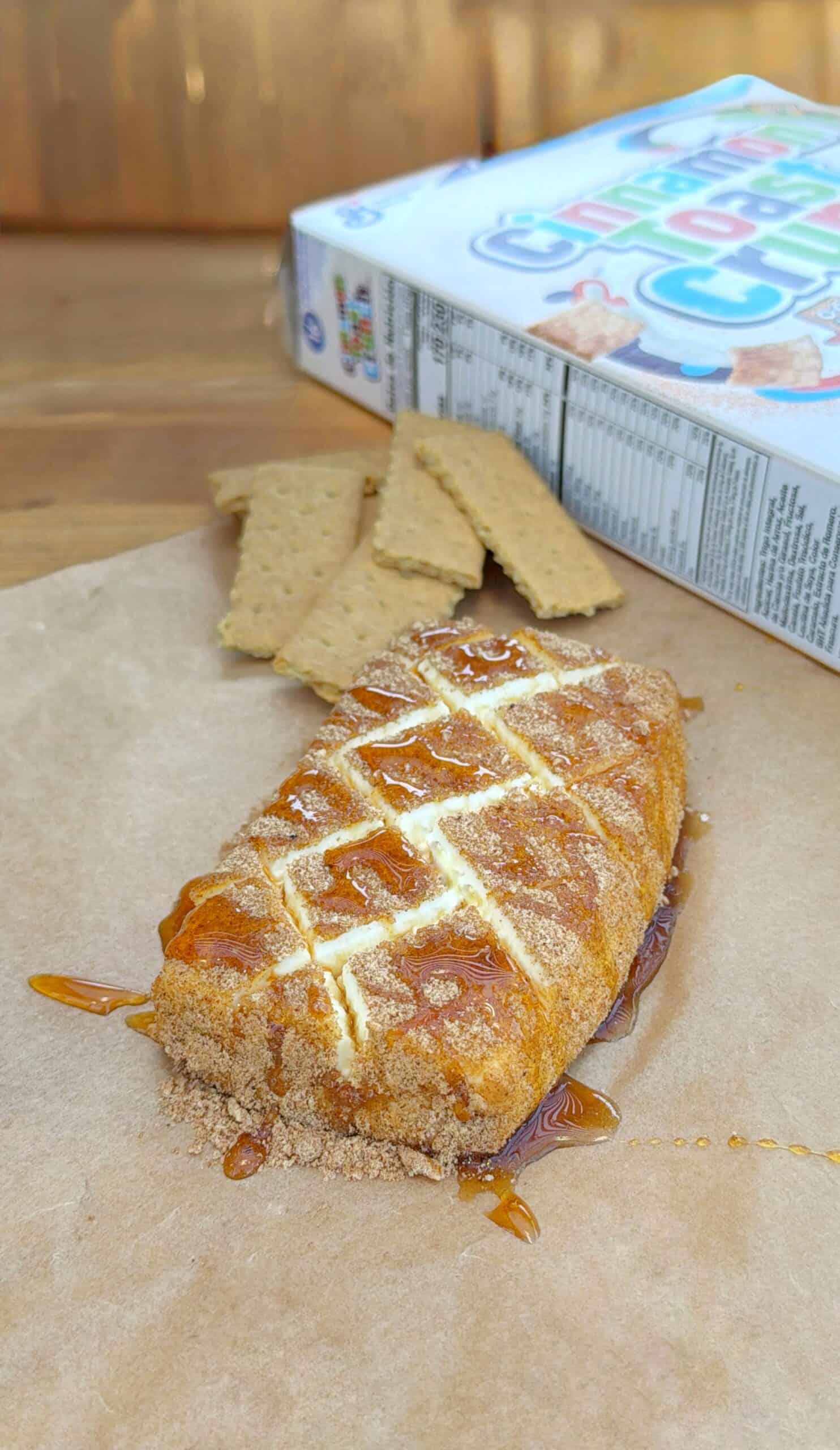 A golden-brown pastry topped with white icing and caramel sauce sits on parchment paper, with graham cracker pieces and a partially visible cereal box in the background.