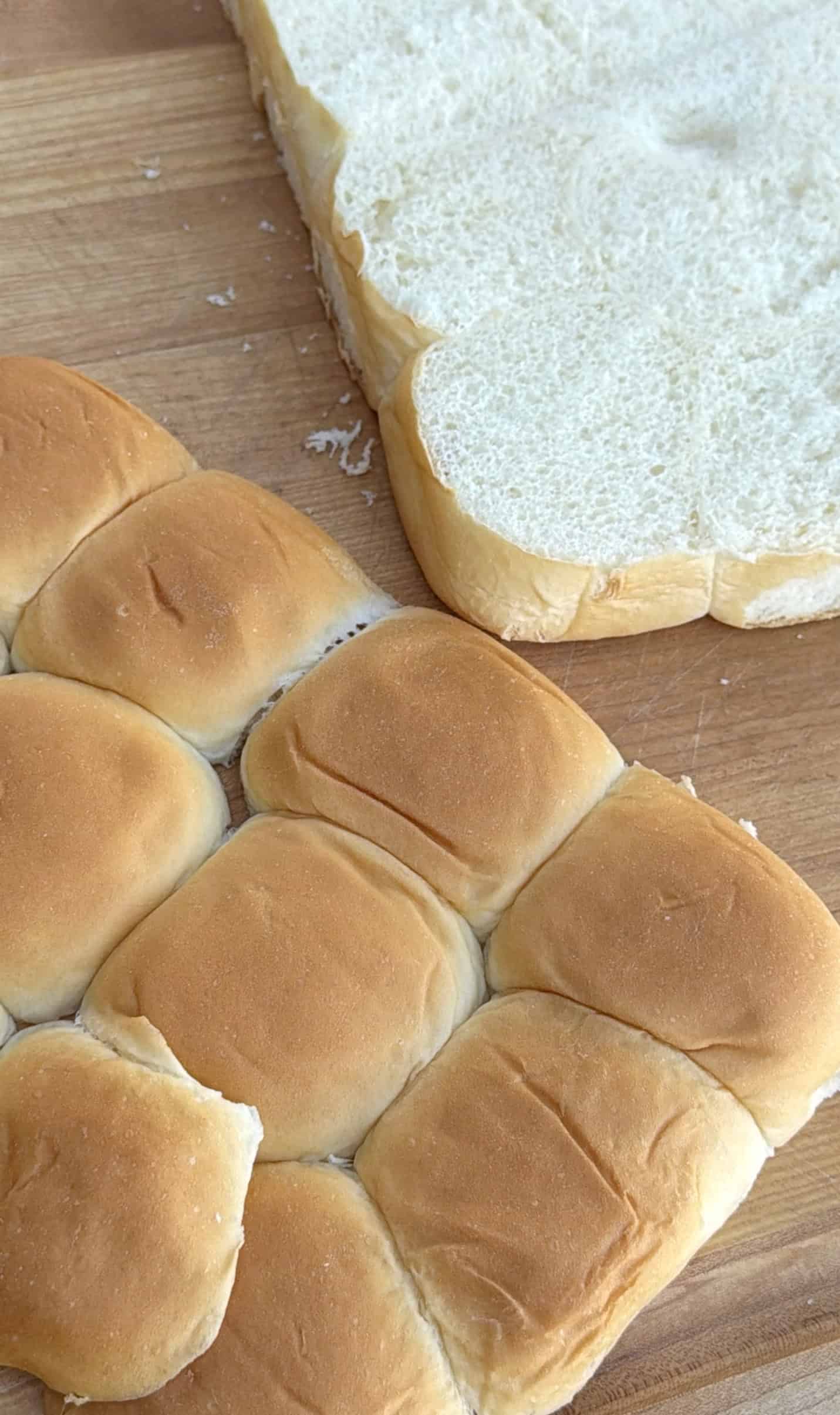 A sheet of soft, golden-brown dinner rolls sits next to its matching top half on a wooden cutting board, showing the fluffy white interior of the bread. For the recipe: Hawaiian Roll Pizza Bread