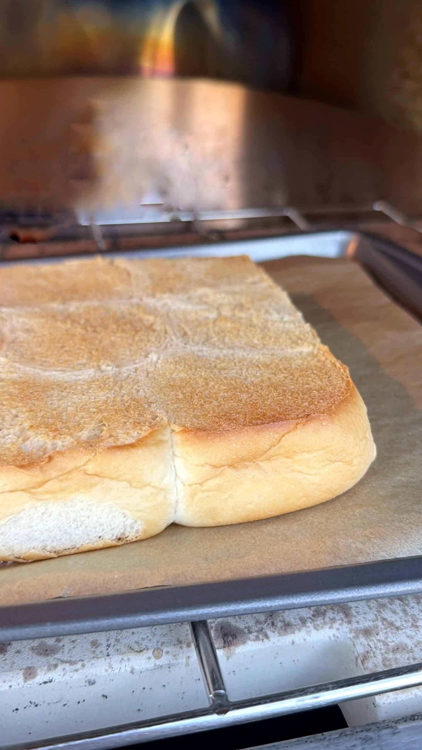 A close-up of a golden-brown loaf of bread on a parchment-lined baking tray, with an oven rack and blurred background visible.