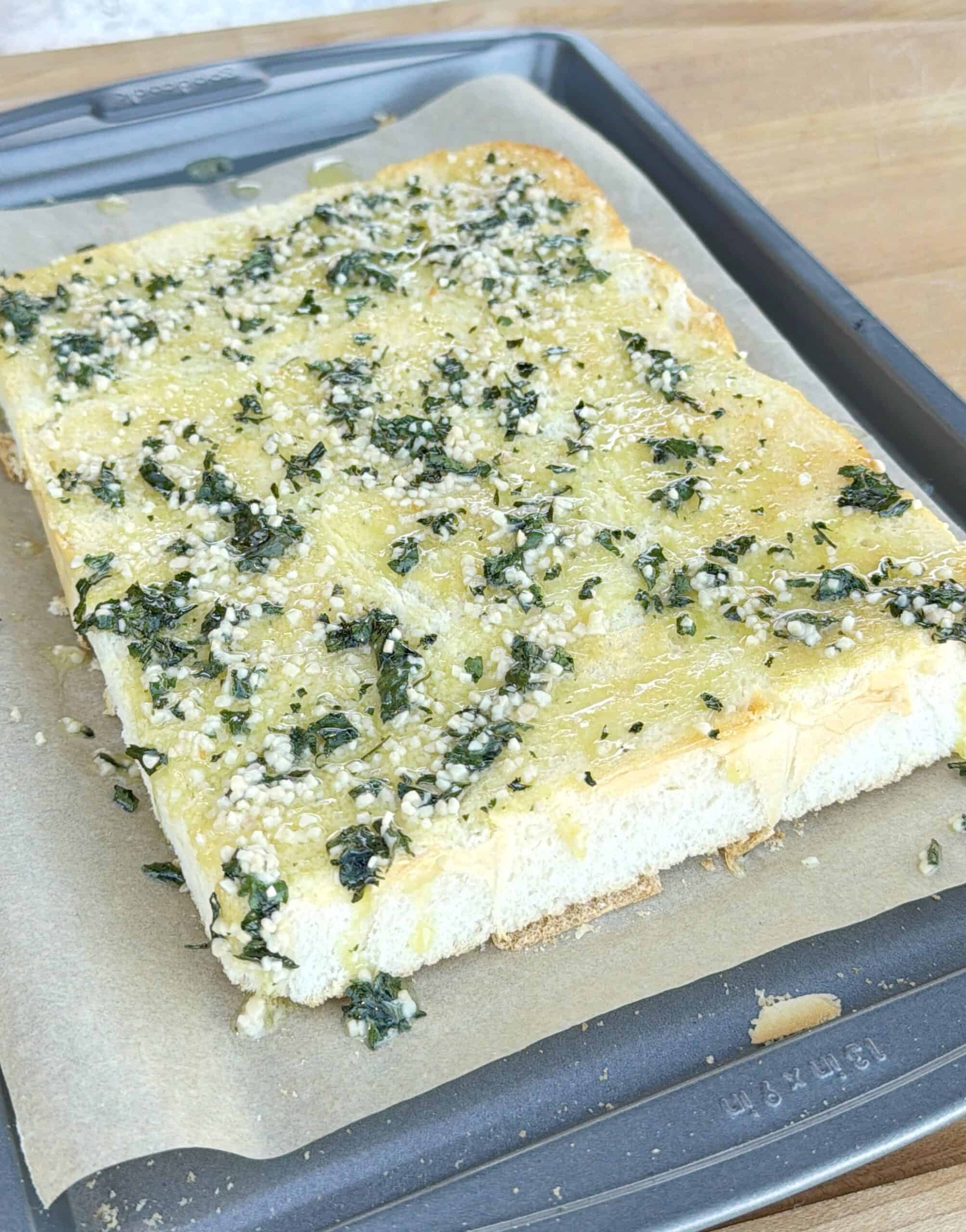A rectangular piece of garlic bread topped with chopped herbs and grated cheese sits on parchment paper on a baking tray. The bread looks toasted and slightly golden on the edges.