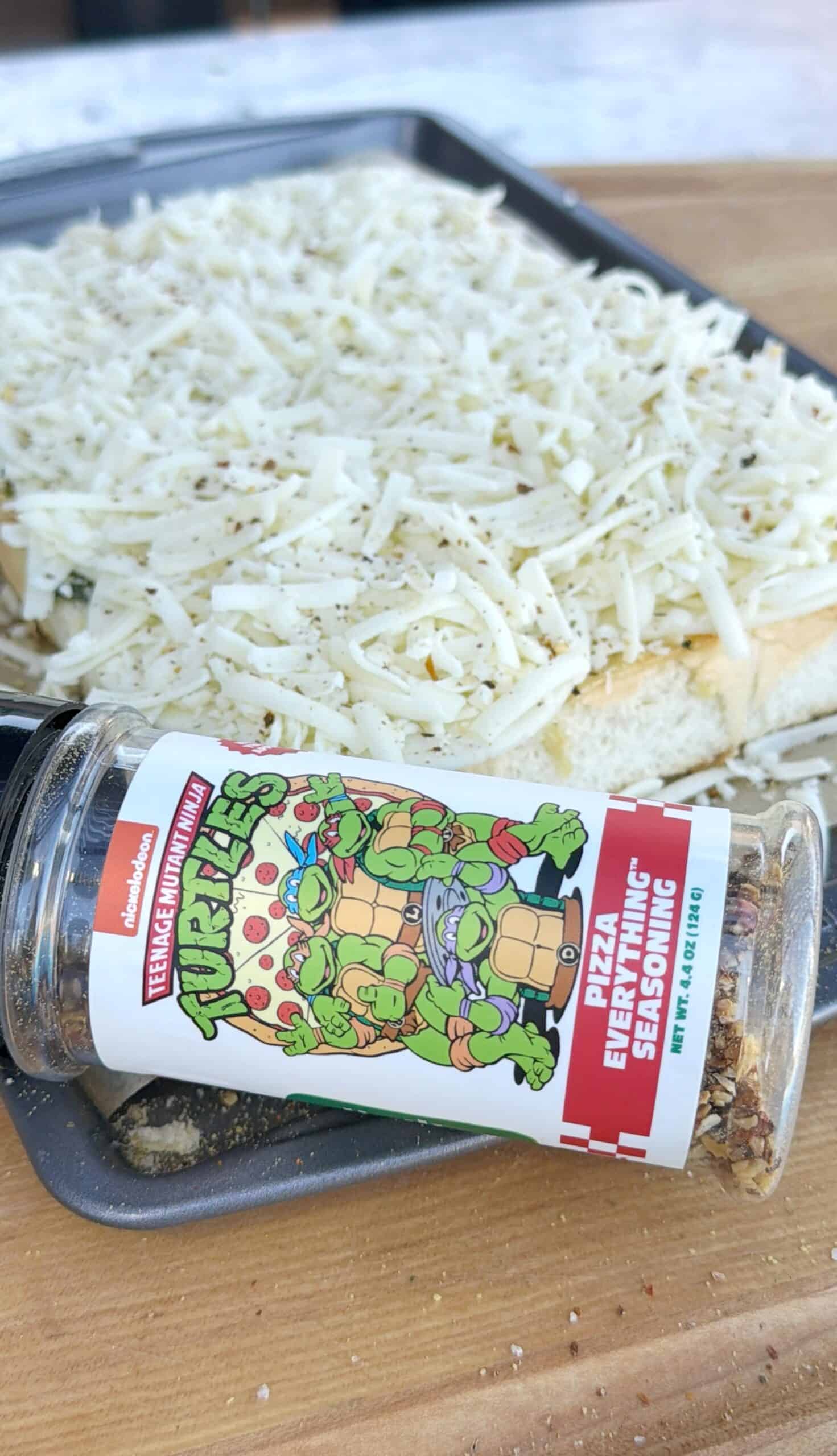 A jar of Teenage Mutant Ninja Turtles Pizza Everything seasoning lies on its side in front of a tray of bread covered with shredded cheese and spices, ready to be baked.