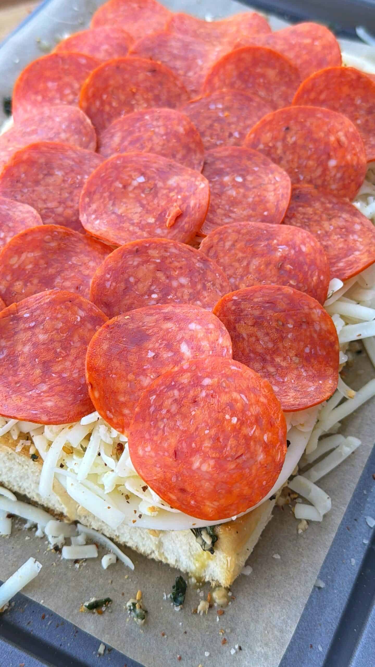 A close-up of a rectangular piece of bread topped with shredded cheese, herbs, and a generous layer of overlapping pepperoni slices on a baking tray.