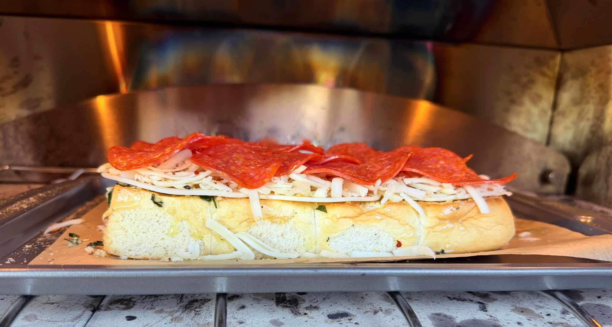 A sub loaf topped with mozzarella cheese and pepperoni slices sits on a metal tray inside an oven, about to be baked.