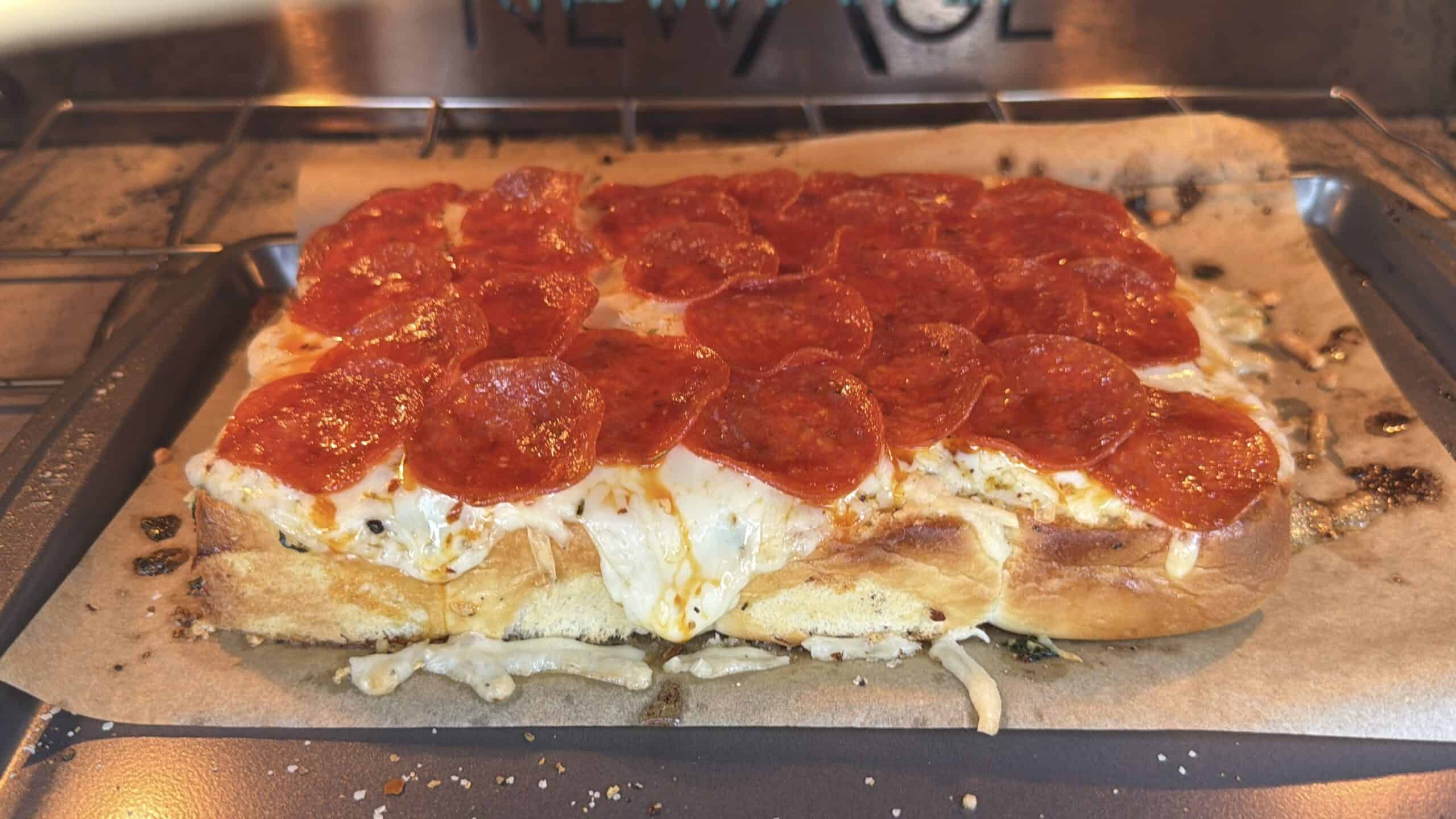A homemade rectangular pepperoni pizza with melted cheese sits on parchment paper in an oven, with the cheese bubbling and some edges slightly browned.