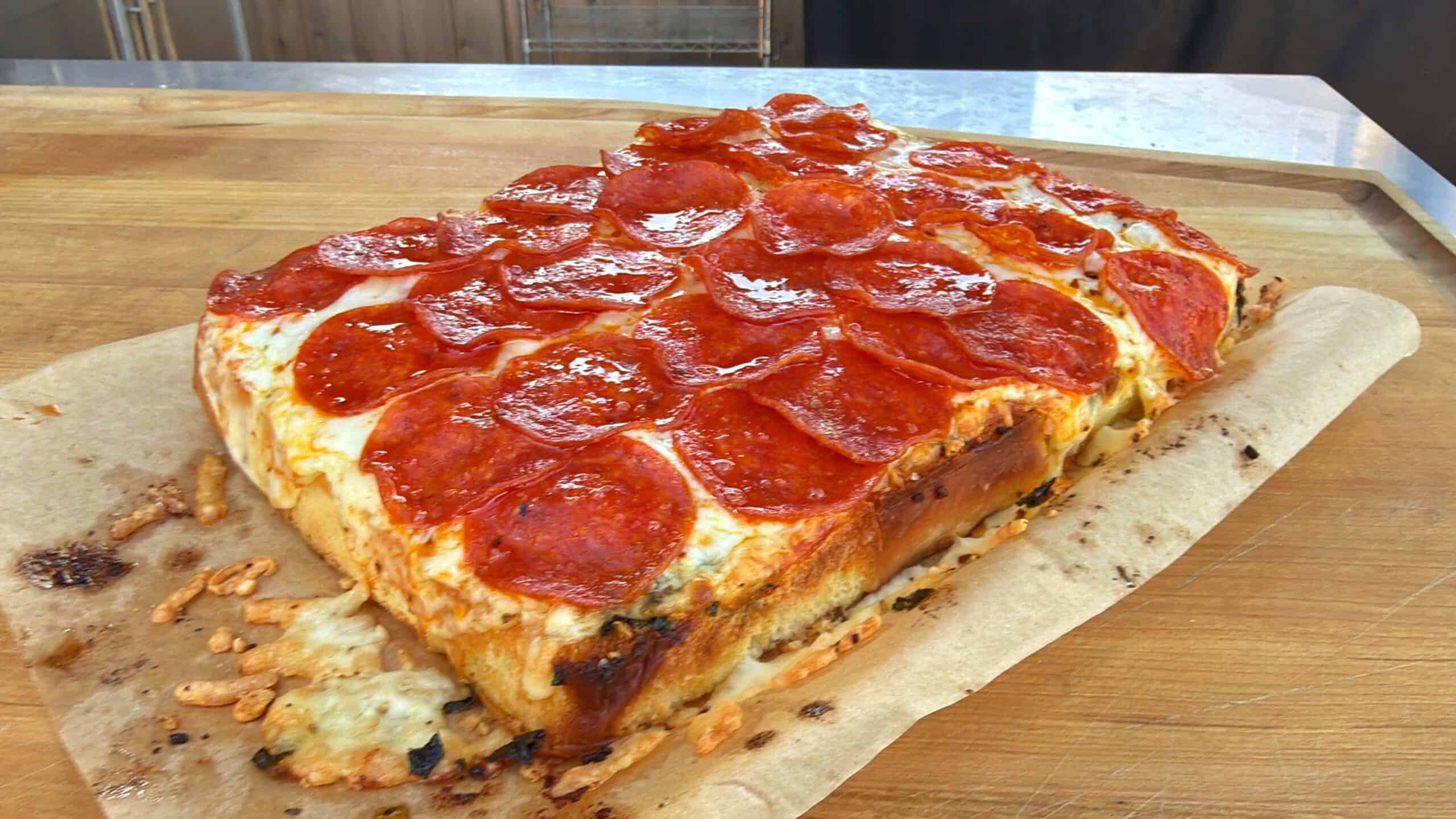 A rectangular, thick-crust pizza topped with melted cheese and pepperoni slices sits on a sheet of parchment paper on a wooden surface.