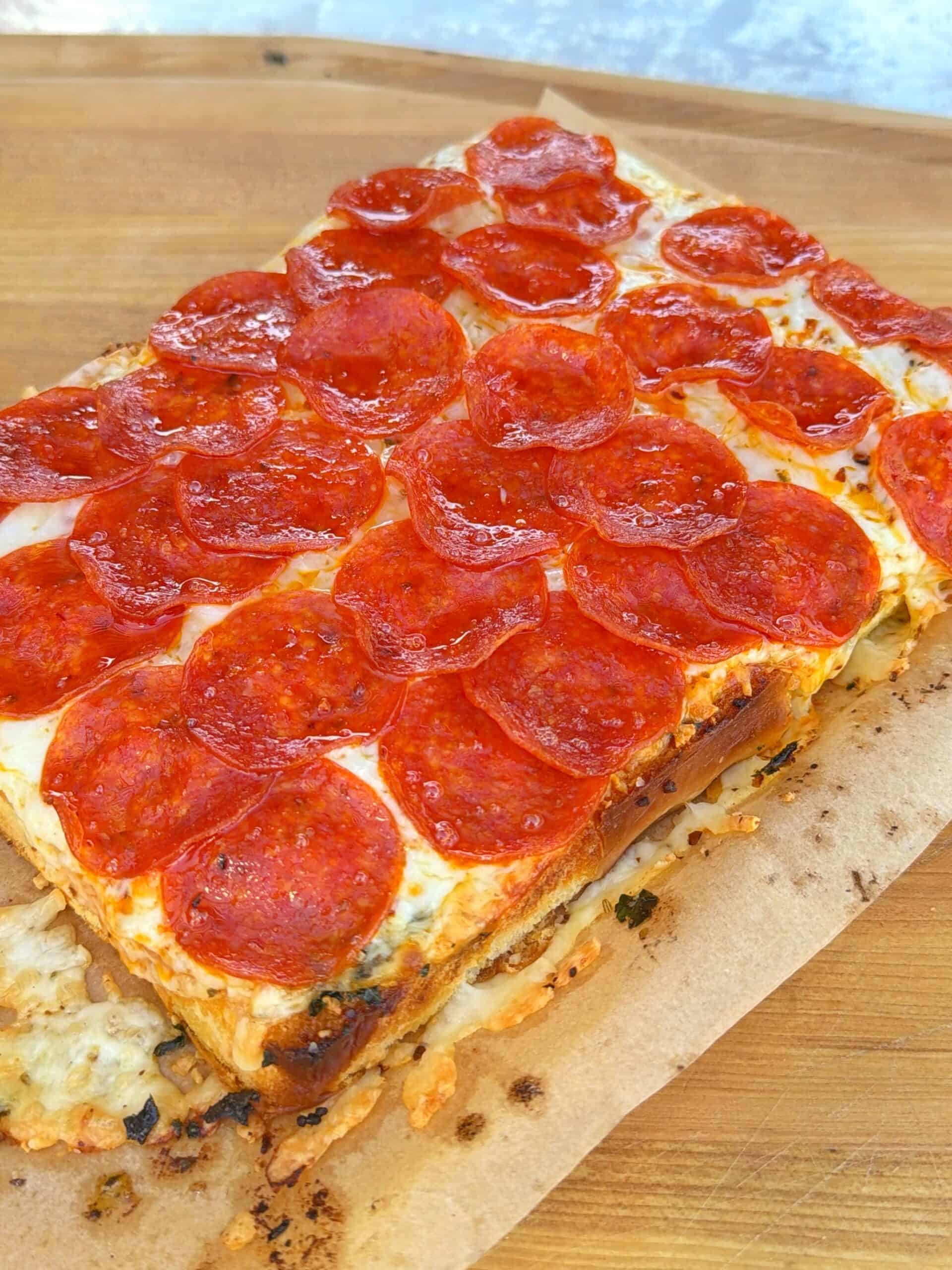 A rectangular pepperoni pizza with a thick, golden crust sits on parchment paper atop a wooden surface. The pizza is generously topped with pepperoni slices and melted cheese.