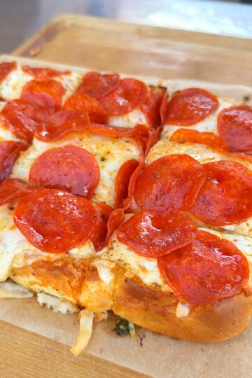 Close-up of a rectangular pepperoni pizza with melted cheese, cut into square pieces, resting on a wooden cutting board. The crust is thick and golden brown, and the pepperoni slices are shiny and slightly crisp.