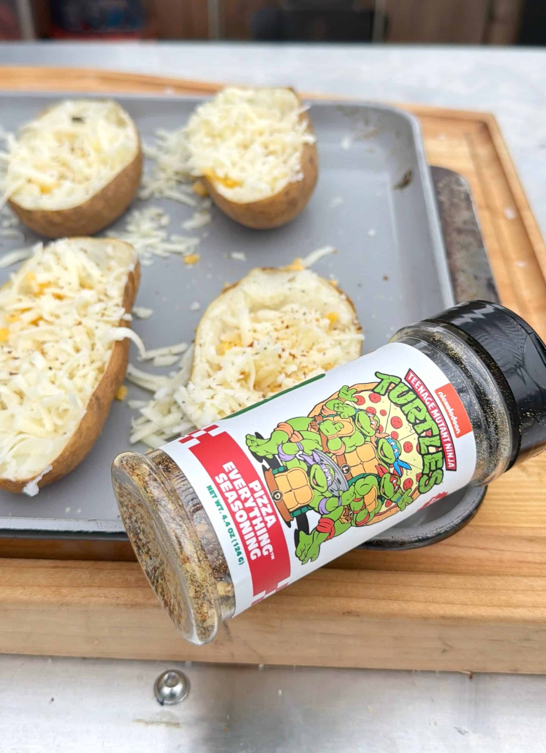 A bottle of Teenage Mutant Ninja Turtles-themed Pizza Everything Seasoning lies on its side in front of a baking tray with halved potatoes topped with shredded cheese and seasoning.
