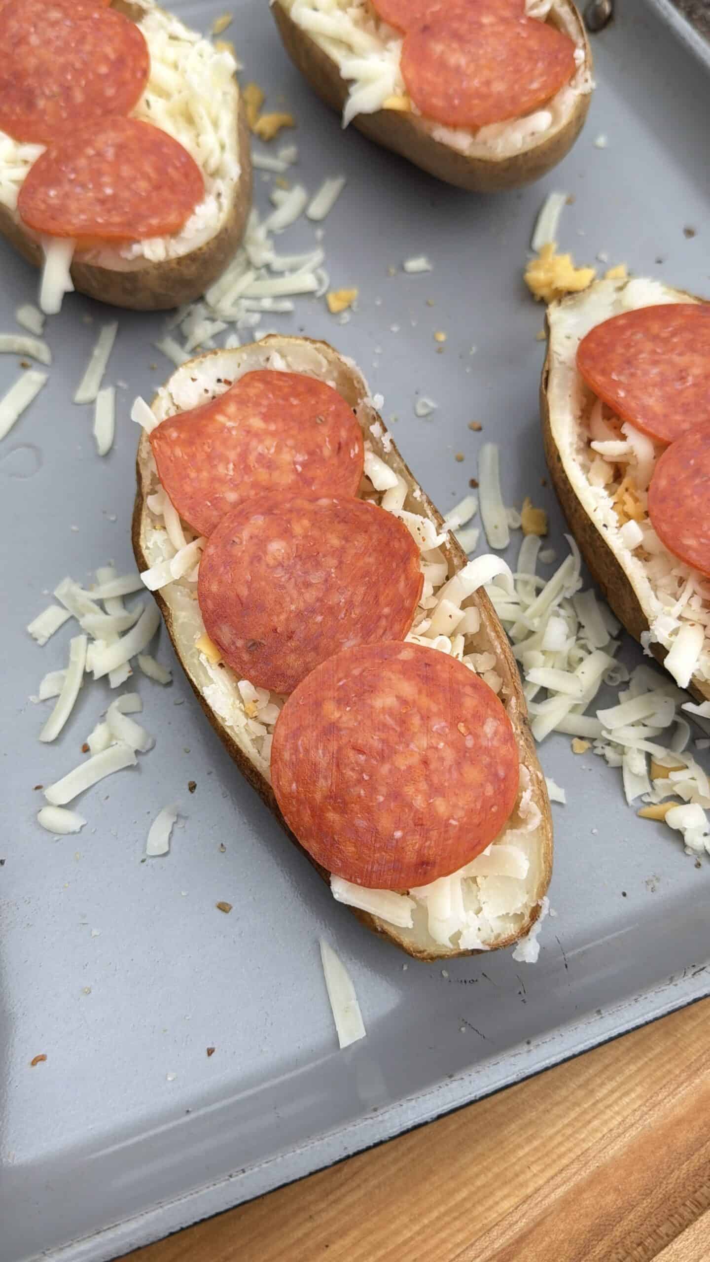 Pizza Potato skins on a baking tray topped with shredded mozzarella cheese and slices of pepperoni, with some cheese scattered on the tray.