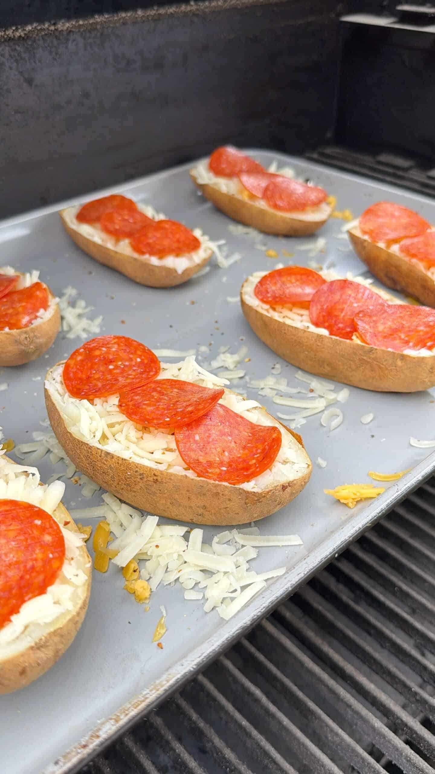Halved potatoes on a baking tray, topped with shredded cheese and pepperoni slices, being cooked on a grill.