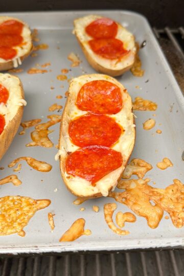 A baking tray holds halved potatoes topped with melted cheese and slices of pepperoni, resembling mini pizza boats. Some cheese is browned and melted on the tray around the potatoes.