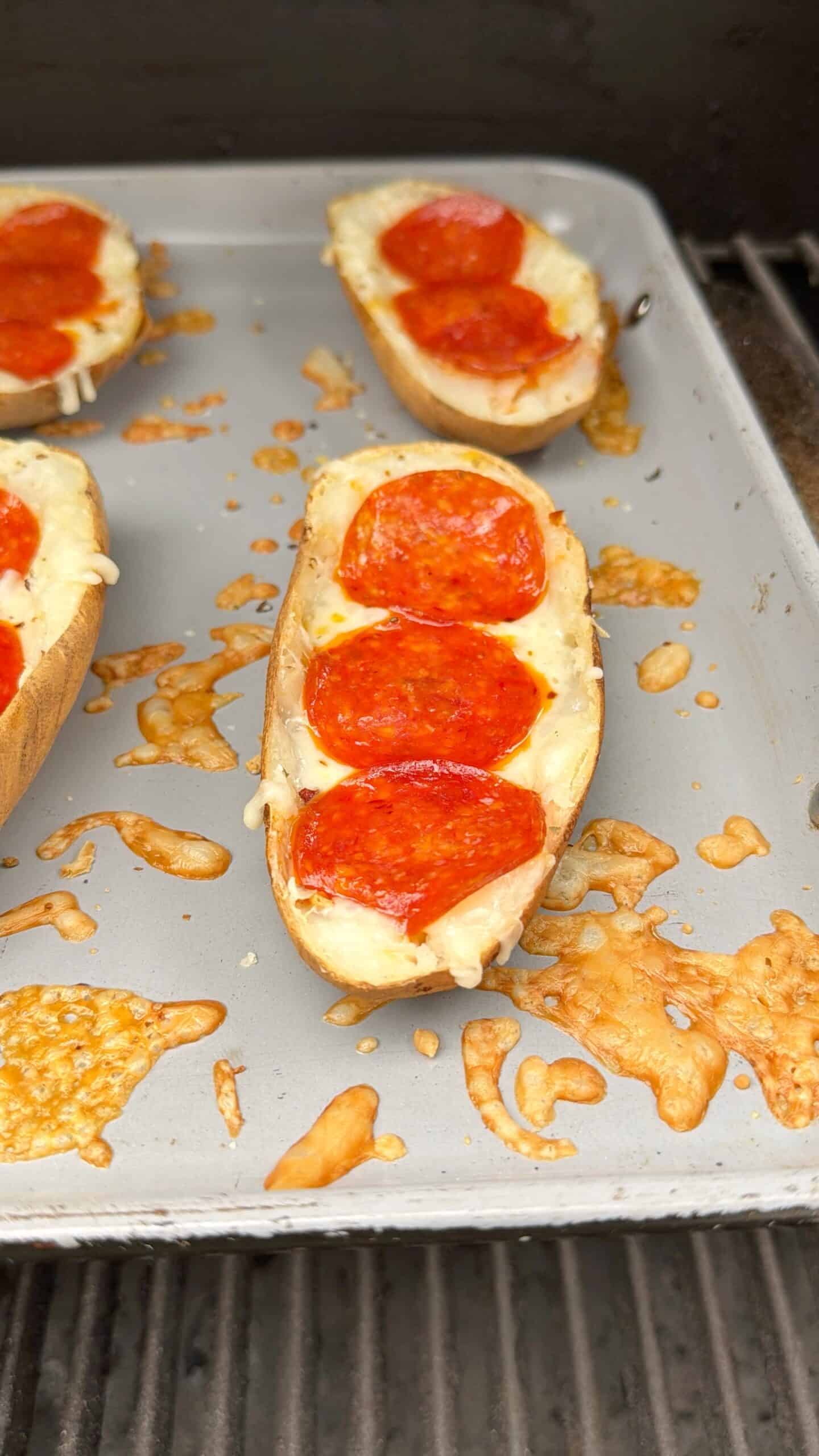 A baking tray holds halved potatoes topped with melted cheese and slices of pepperoni, resembling mini pizza boats. Some cheese is browned and melted on the tray around the potatoes.