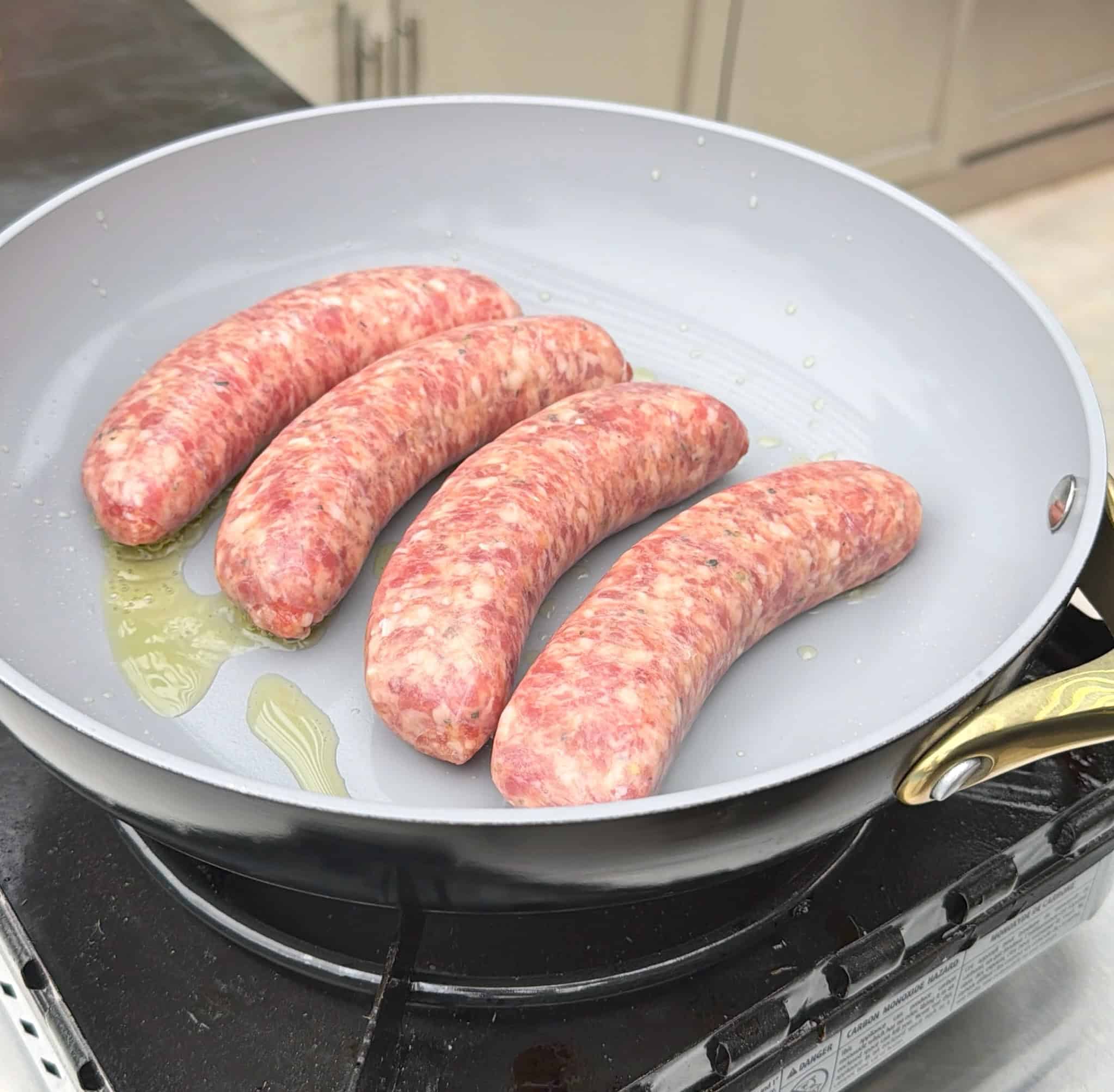 Four raw sausages are cooking in a lightly oiled frying pan on a stovetop. The sausages are arranged side by side and have not yet browned. The kitchen countertop and cabinets are visible in the background.