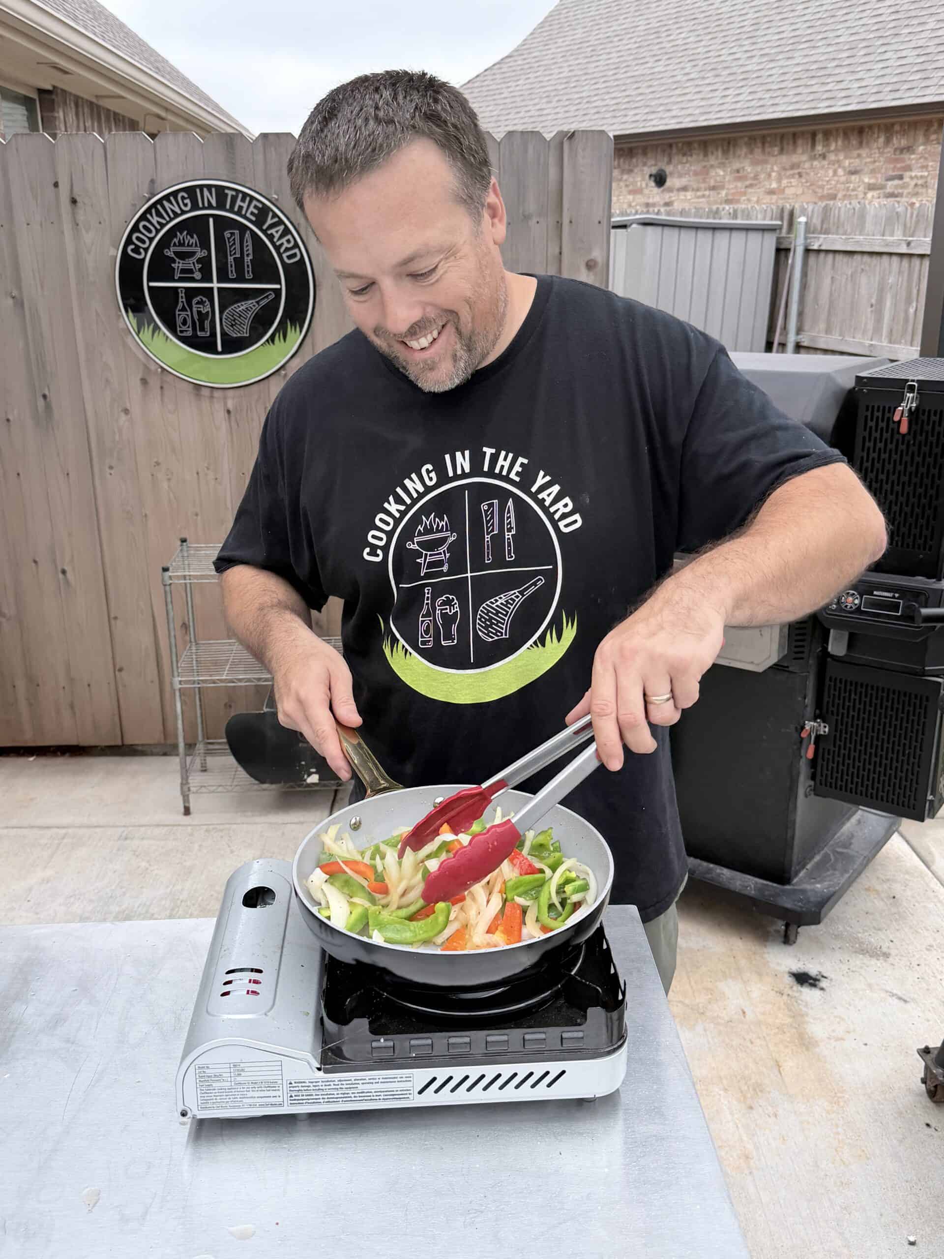 A smiling man cooks colorful vegetables in a frying pan on a portable stove outdoors. He is using tongs and wearing a black Cooking in the Yard t-shirt. A wooden fence and a sign are visible in the background.