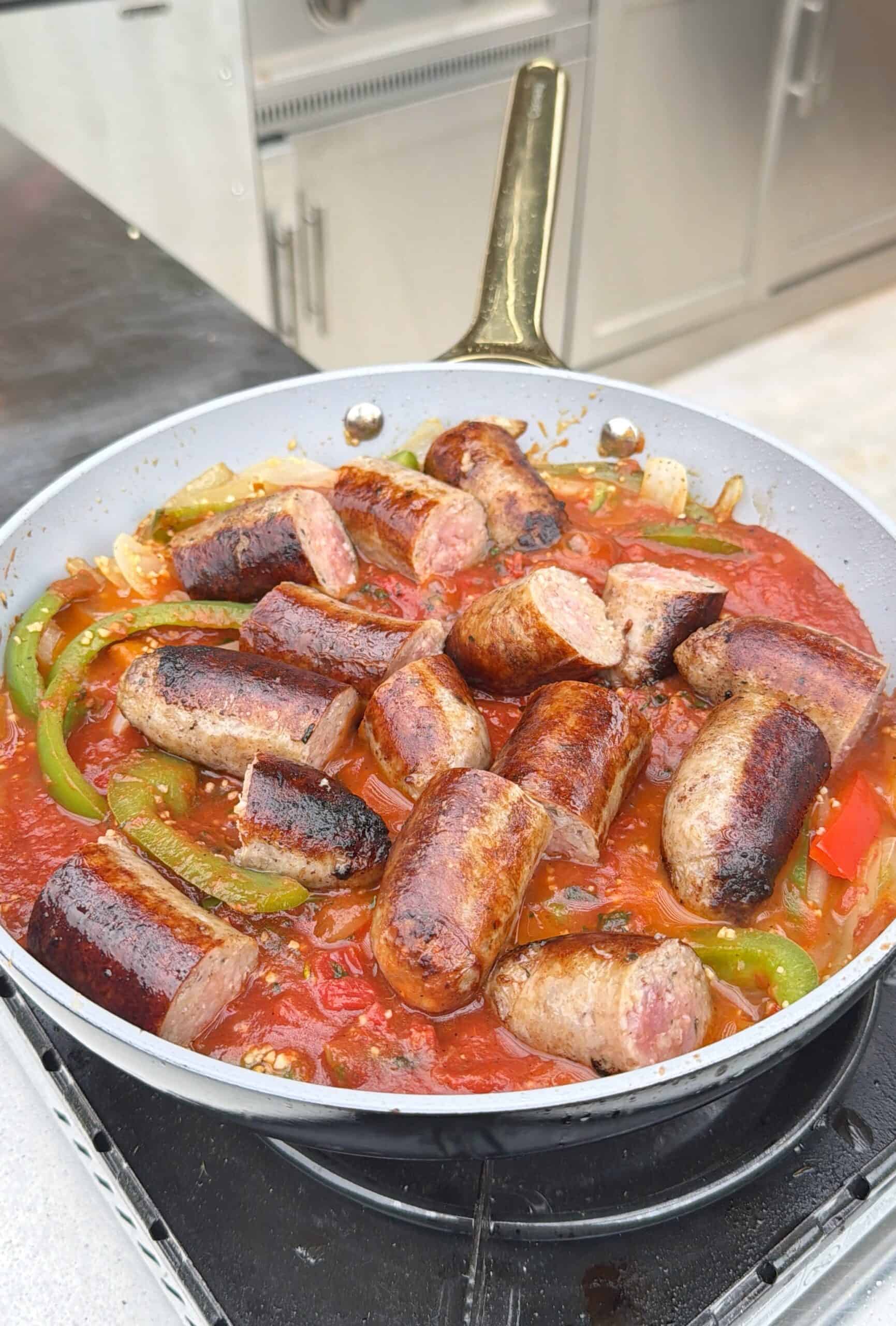 A skillet filled with browned sausage pieces simmering in a tomato sauce with sliced green peppers and onions, cooking on an outdoor stovetop.