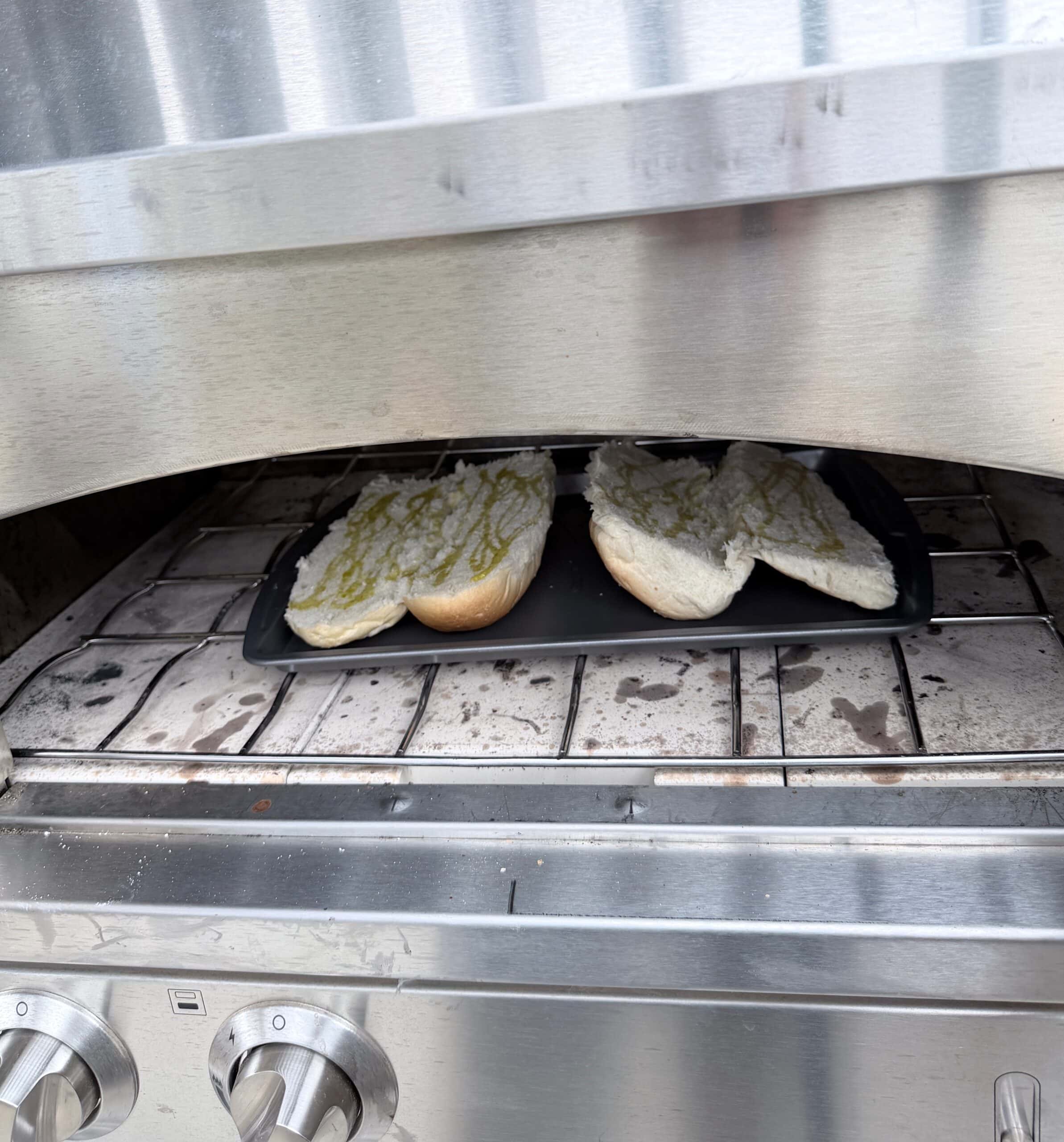 Two open sandwich rolls with a spread, likely butter or oil, are toasting on a tray inside a stainless steel outdoor oven or grill. The oven has a tile base and control knobs visible in the foreground.