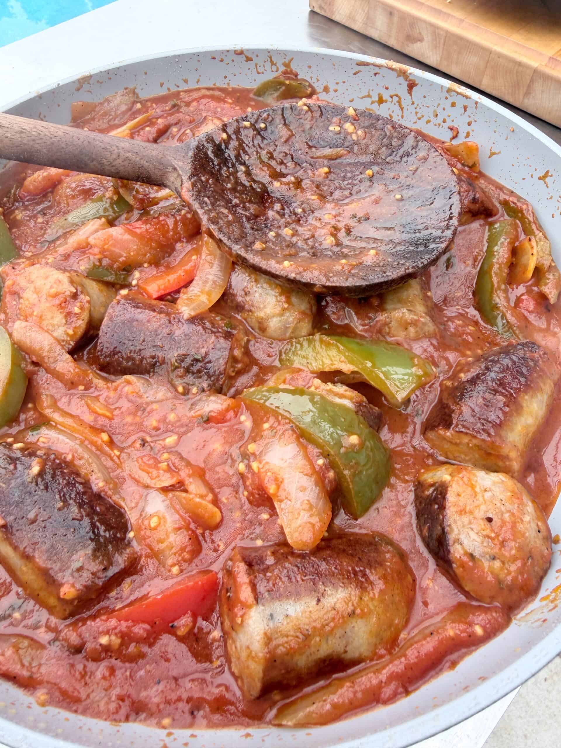 Sausage pieces with bell peppers and onions simmer in a rich tomato sauce, stirred with a wooden spoon in a large pan.